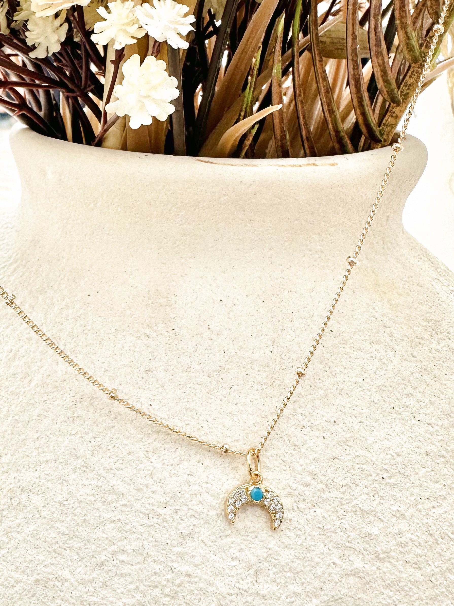 Gold crescent moon necklace with turquoise centre and cubic zirconia displayed on textured vase with dried florals