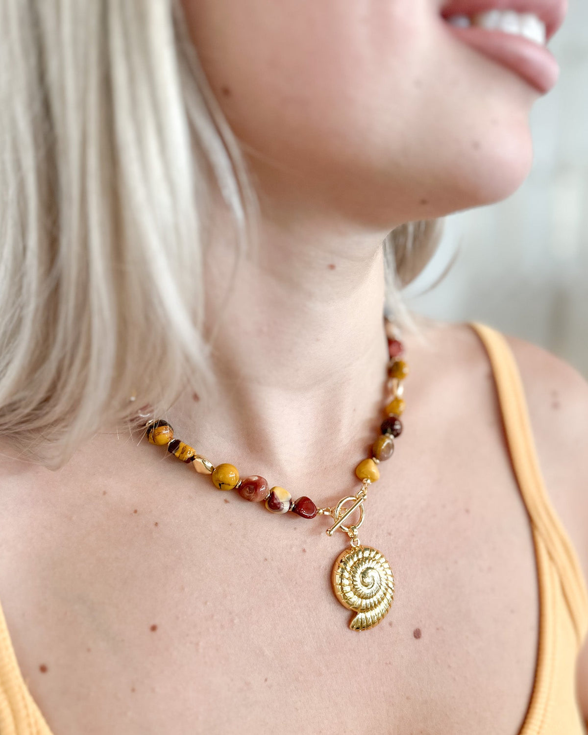 A close-up lifestyle shot of a woman wearing a handmade beaded necklace with a gold spiral shell pendant. The necklace features a mix of amber, brown, and red beads with a gold toggle clasp.