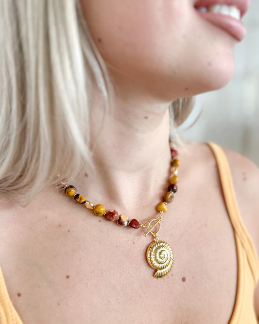 A close-up lifestyle shot of a woman wearing a handmade beaded necklace with a gold spiral shell pendant. The necklace features a mix of amber, brown, and red beads with a gold toggle clasp.