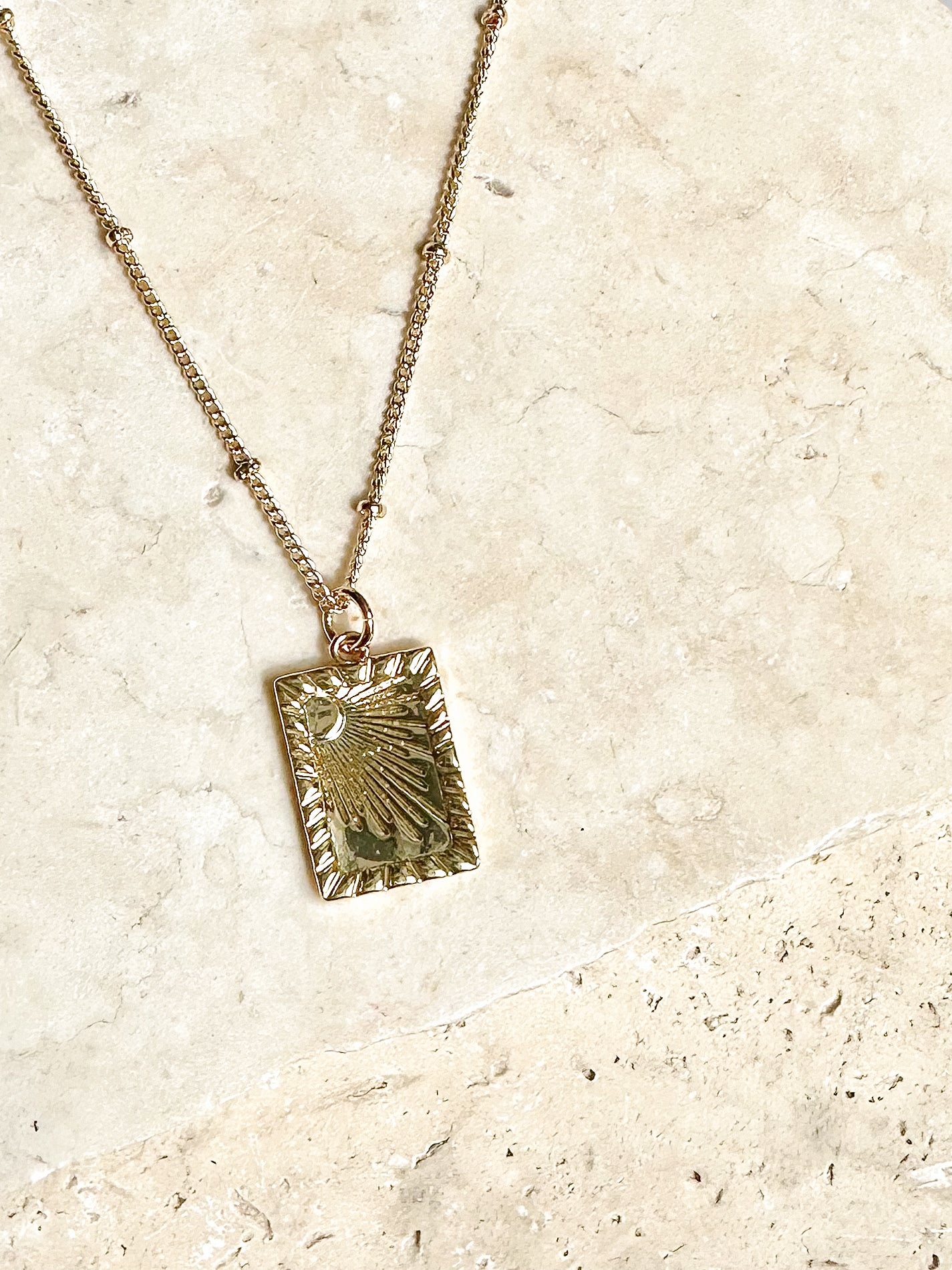 Gold rectangular sunbeam pendant necklace displayed on textured ceramic surface 