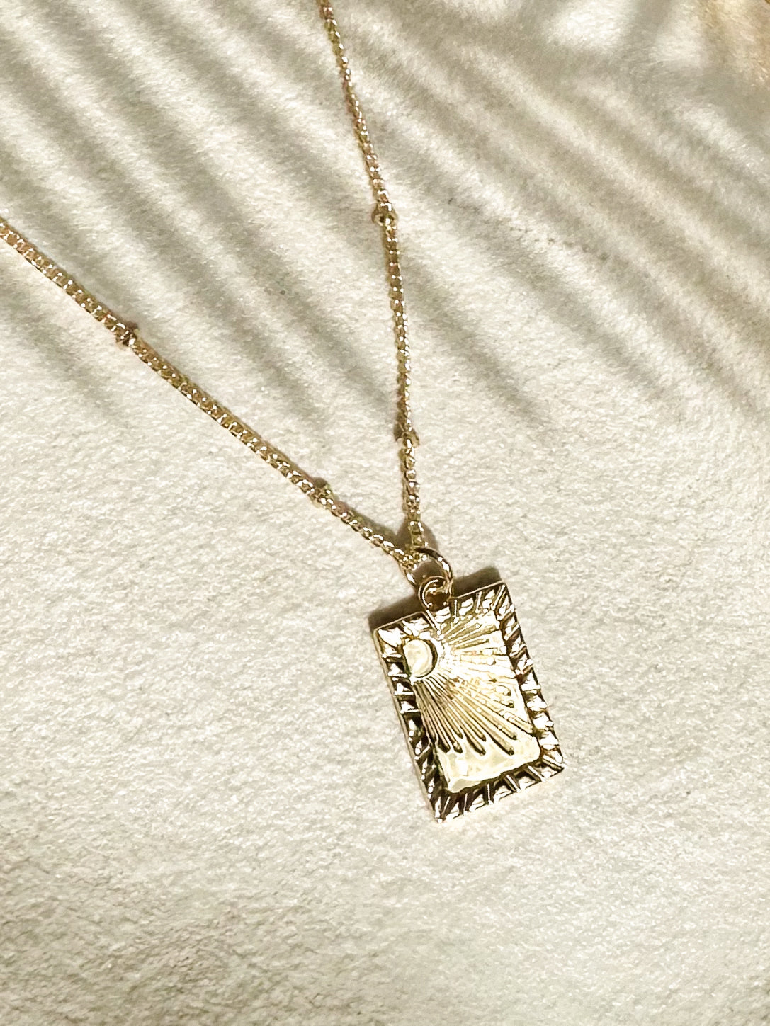 Gold rectangular sunbeam pendant necklace displayed on textured ceramic surface 