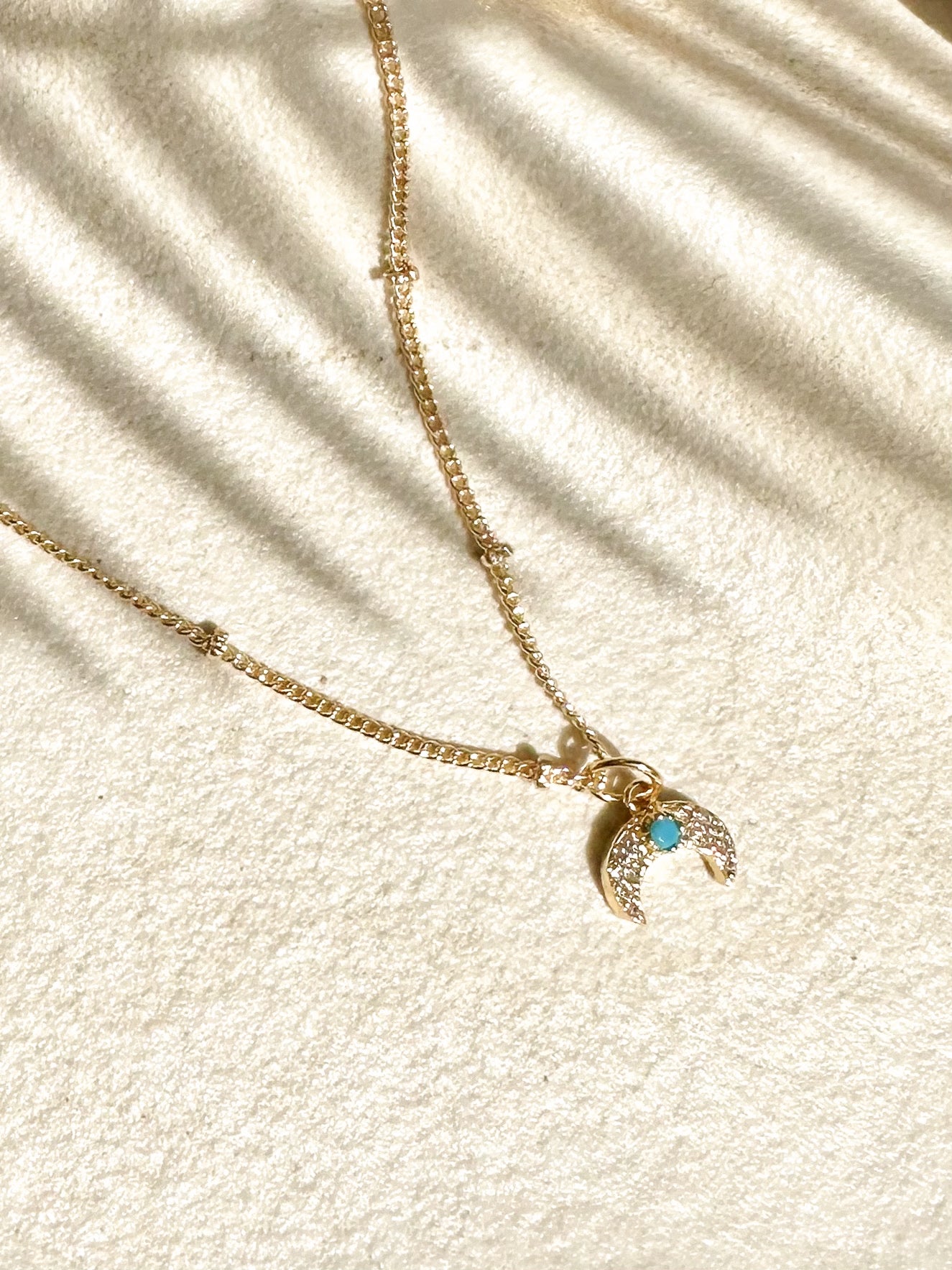 Gold crescent moon necklace with turquoise centre and cubic zirconia displayed on travertine with soft palm leaf shadows