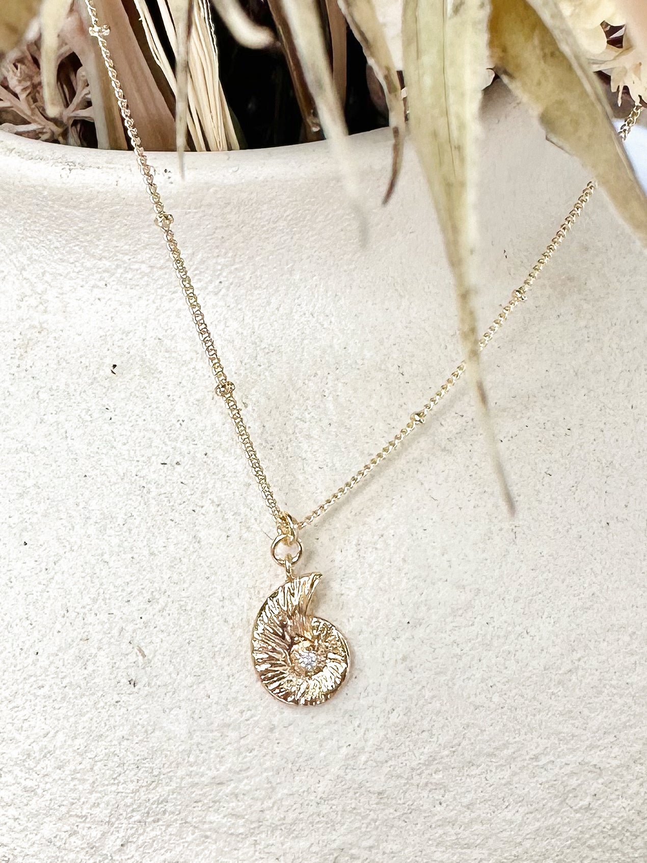 Gold nautilus shell pendant necklace with cubic zirconia centre displayed on textured ceramic vase and dried florals.