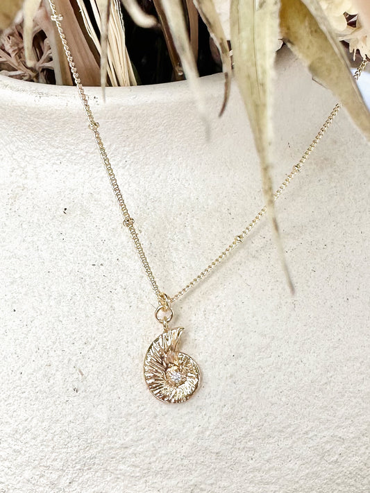 Gold nautilus shell pendant necklace with cubic zirconia centre displayed on textured ceramic vase and dried florals.