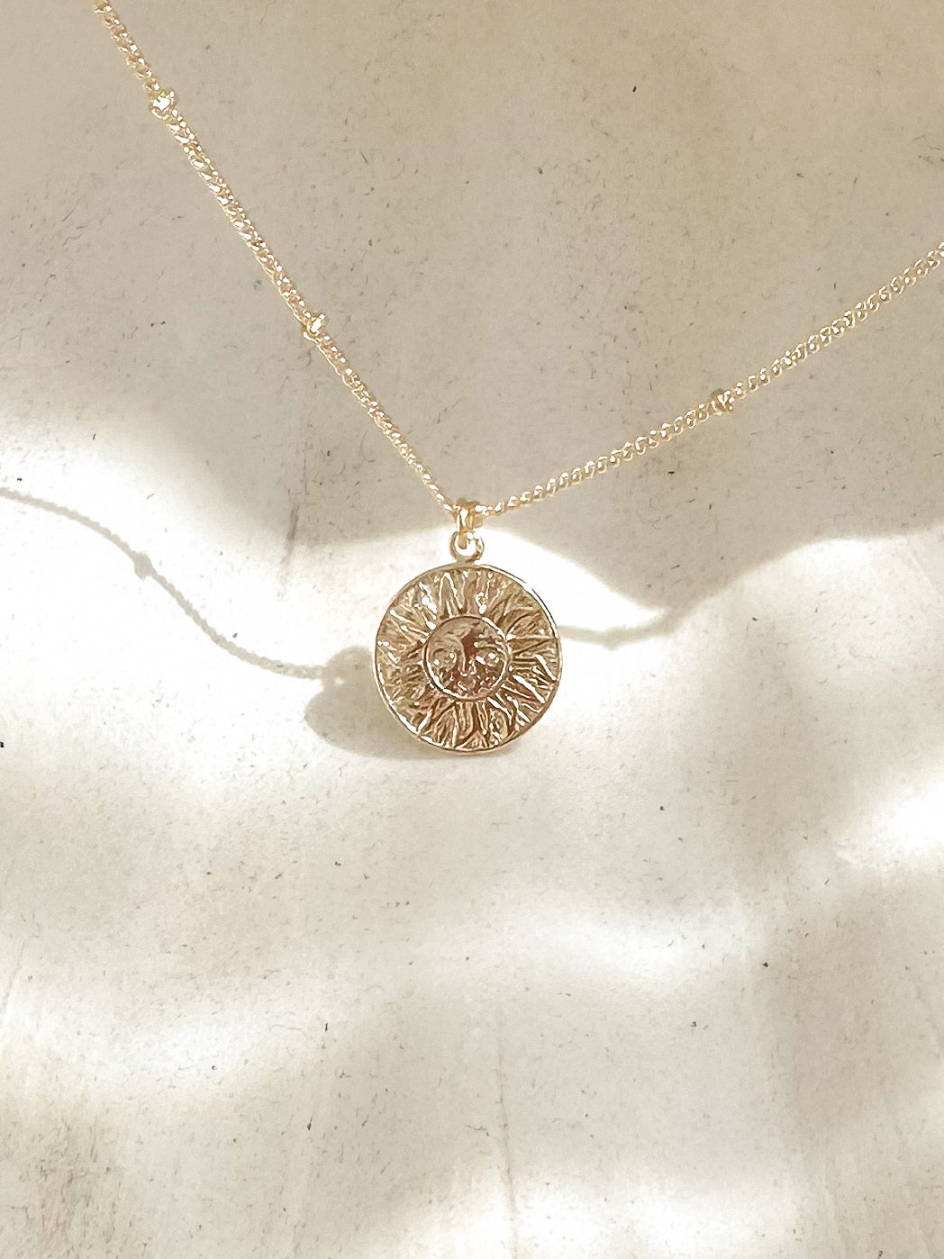 Gold sun pendant necklace displayed on soft textured background with warm sunlight.