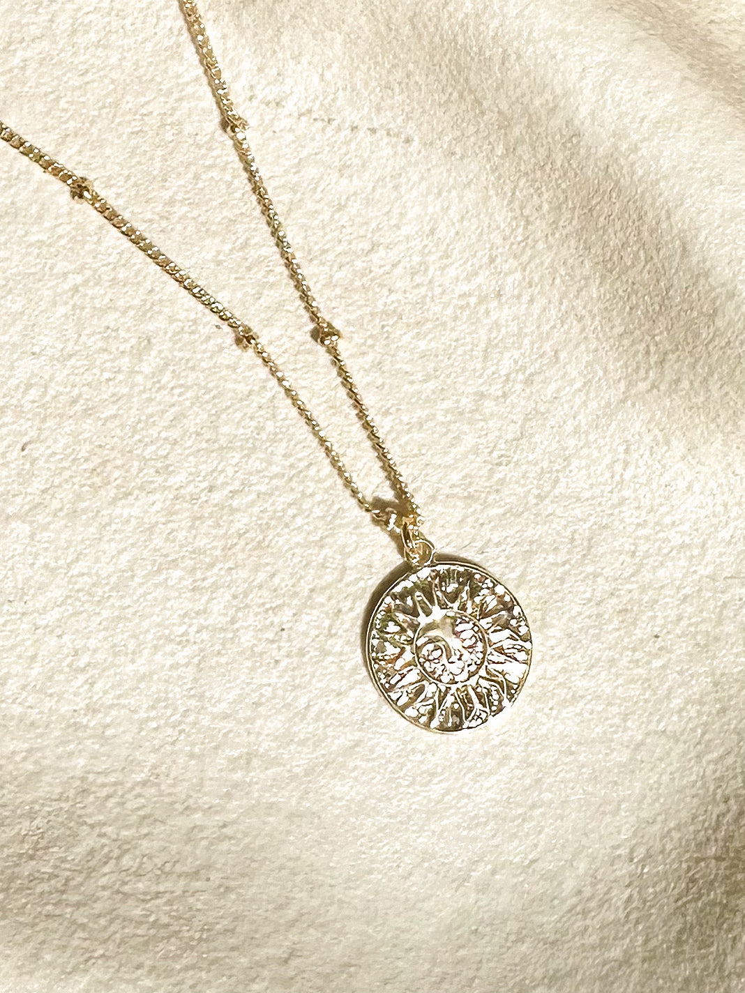 Gold sun pendant necklace displayed on soft textured background with warm sunlight.