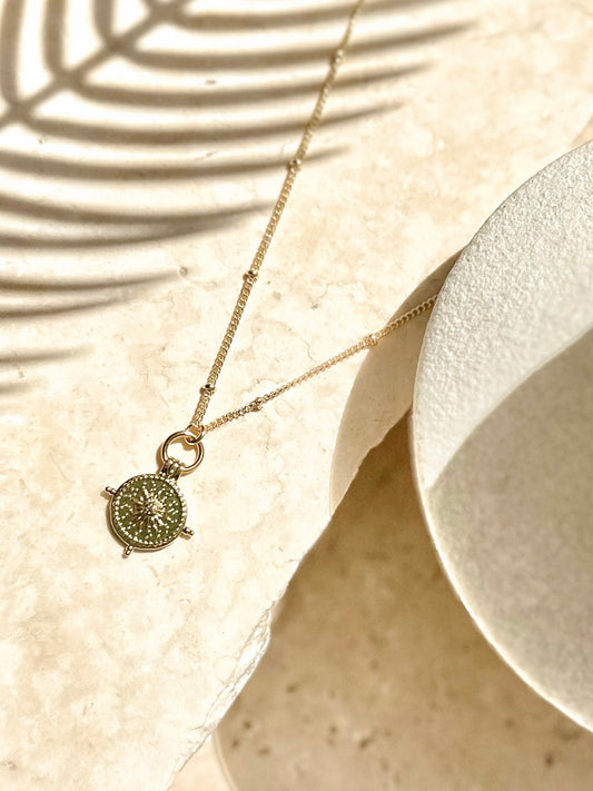 Gold sunburst coin pendant necklace displayed on travertine with soft palm shadows.