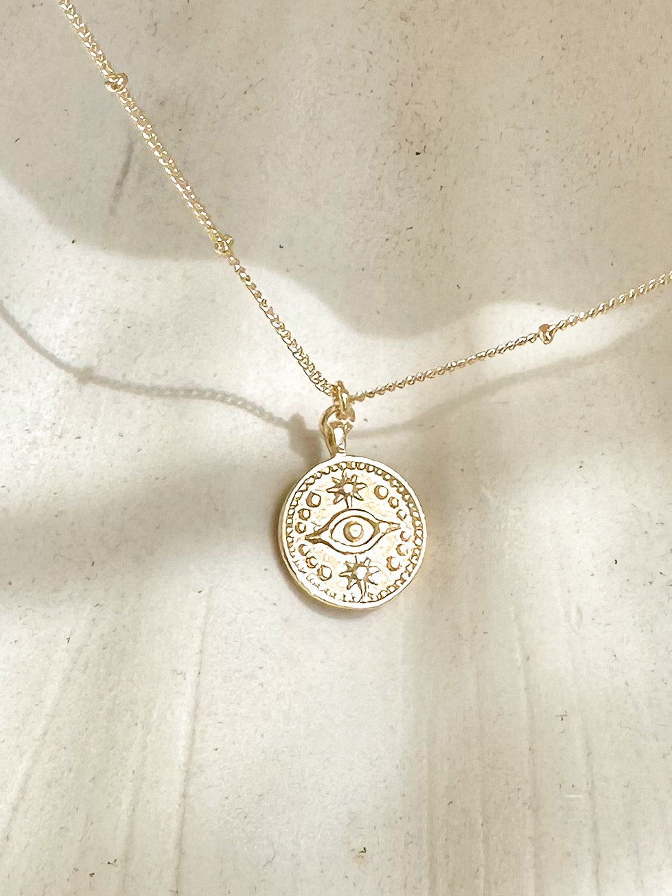 Gold evil eye coin pendant necklace displayed on shell with sunlight casting soft shadows.