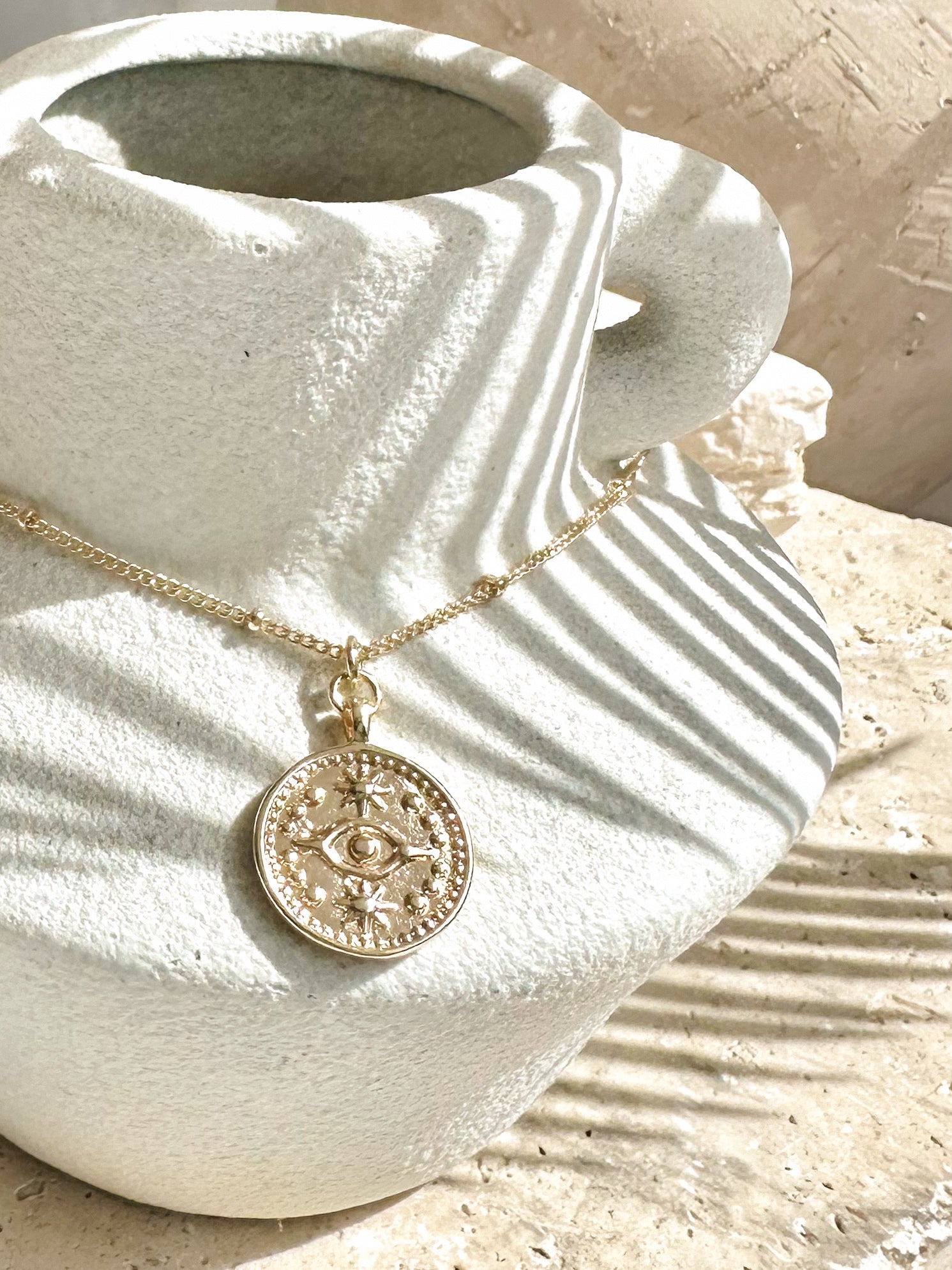 Gold evil eye coin pendant necklace displayed on textured ceramic vase with sunlight casting soft shadows.