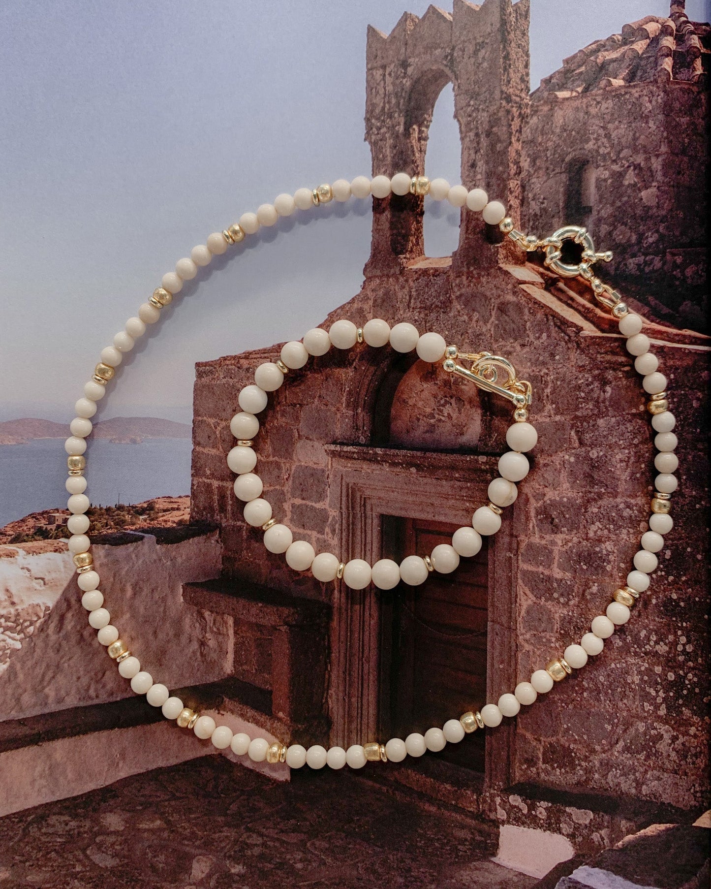 A Riverstone beaded necklace and matching bracelet with gold-toned spacers and clasp, arranged over a scenic wall print of a Mediterranean stone building and sea.