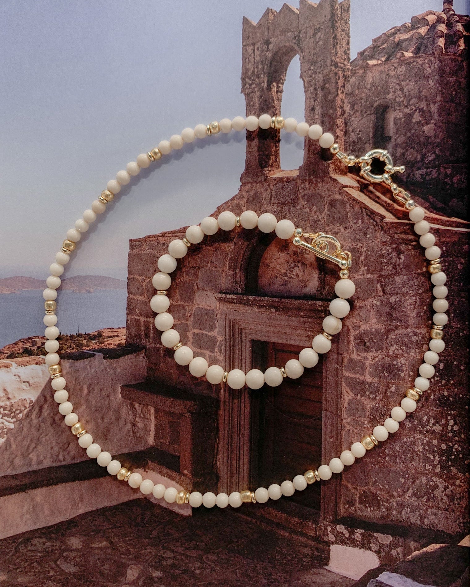 A Riverstone beaded necklace and matching bracelet with gold-toned spacers and clasp, arranged over a scenic wall print of a Mediterranean stone building and sea.