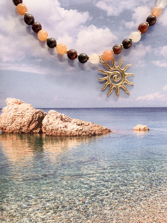 A beaded necklace with a gold sun pendant is laid diagonally on a split background of blue water and a cloudy sky, alongside a small seashell.