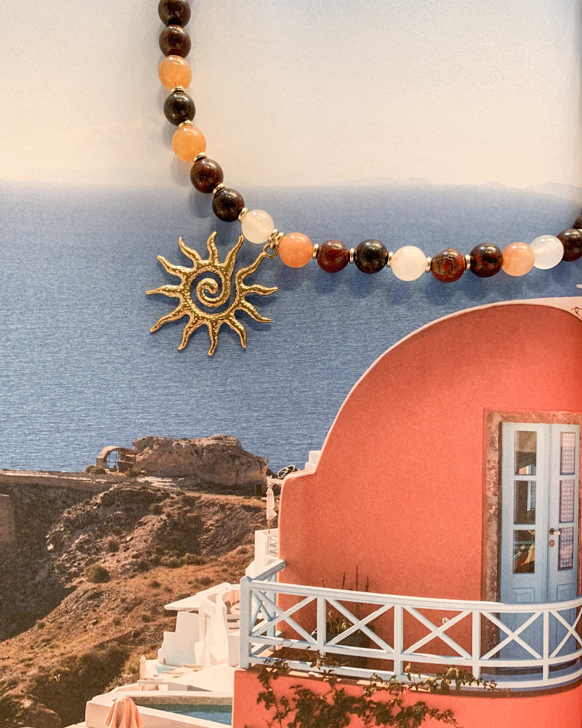 A beaded necklace with a gold sun pendant is draped across a scenic print of Santorini, featuring a pink building, white railing, and blue sea.