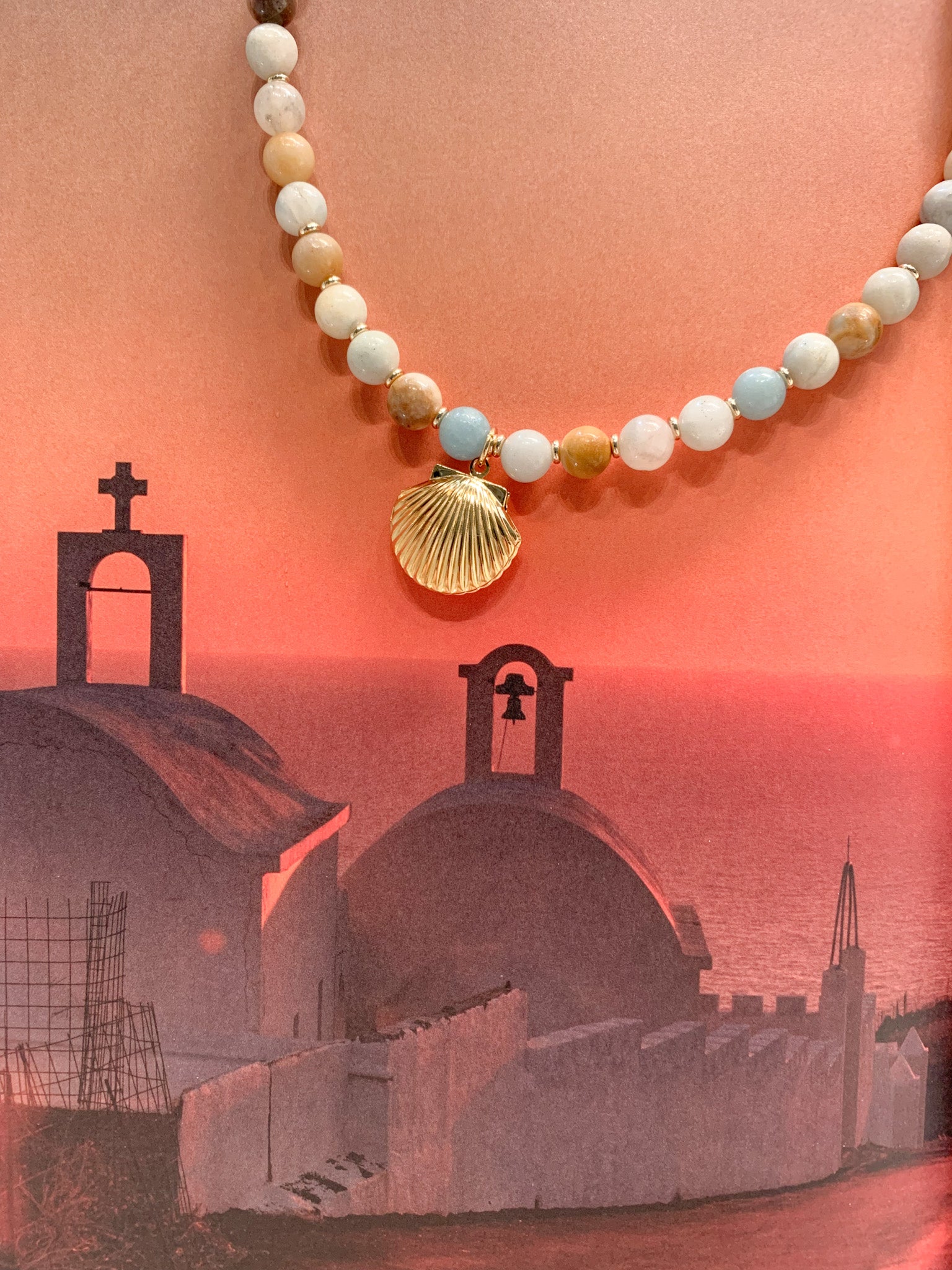 A beaded necklace with a gold seashell pendant, displayed against a warm peach background with a silhouette of a coastal church and sea.