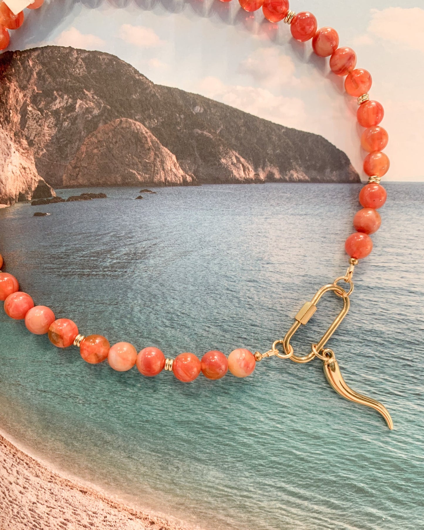 A beaded necklace made of round coral-orange and pinkish-red stones with a gold toggle clasp, laid over a beach-and-sea background photo.