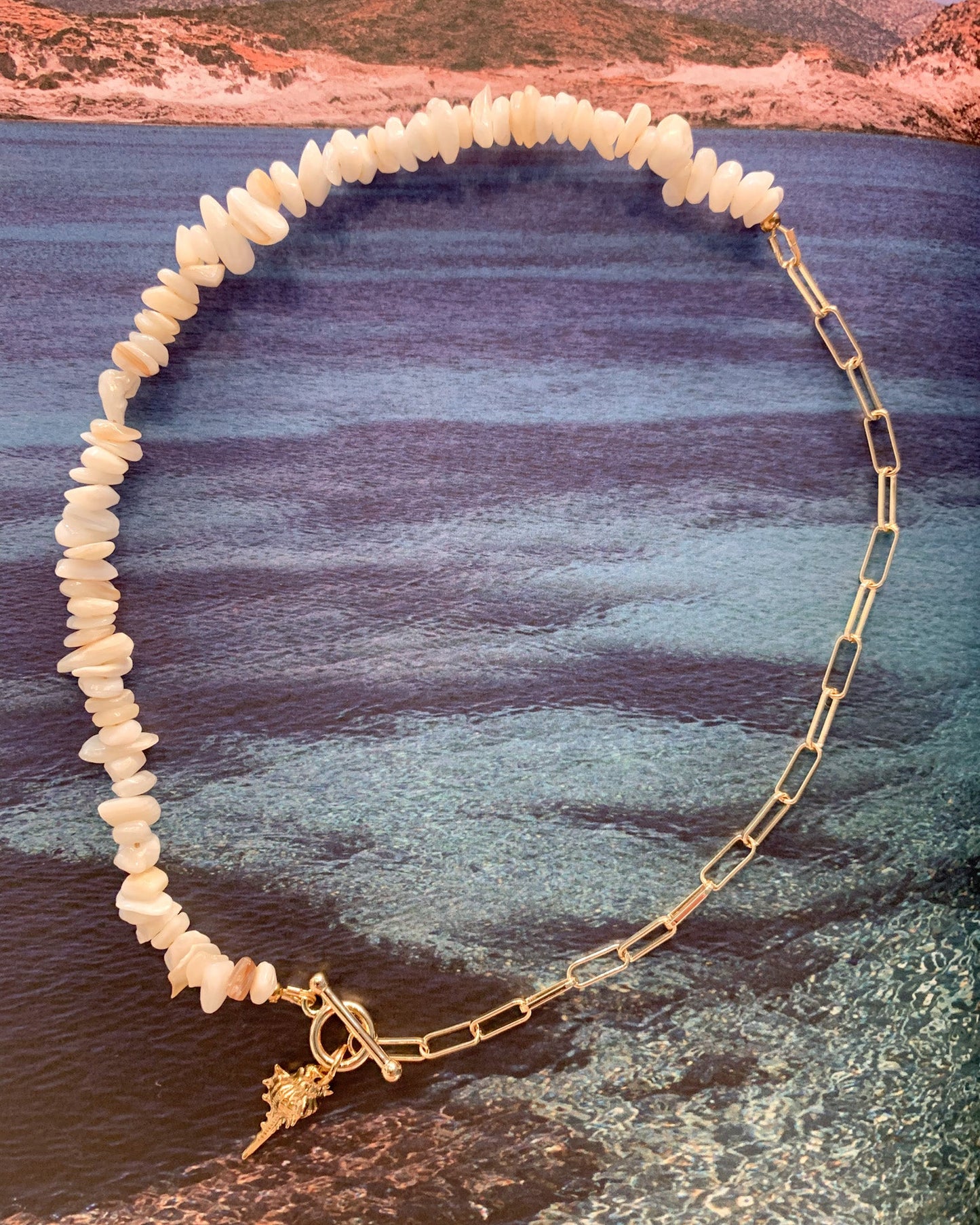 A gold chain necklace with irregular white shell beads and a small gold clasp, arranged in an oval shape over a scenic coastal background of blue water and mountains.