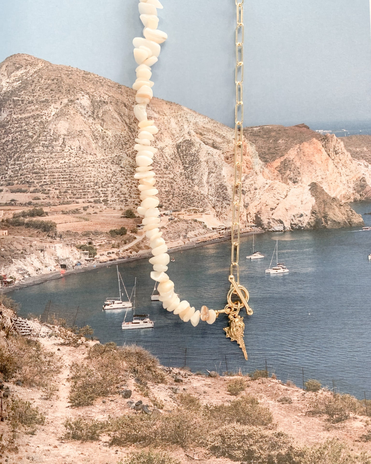 A gold chain necklace draped vertically in the foreground, featuring a large shell-bead strand and a gold cross pendant, set against a scenic coastal background with blue water, rocky cliffs, and sailboats.