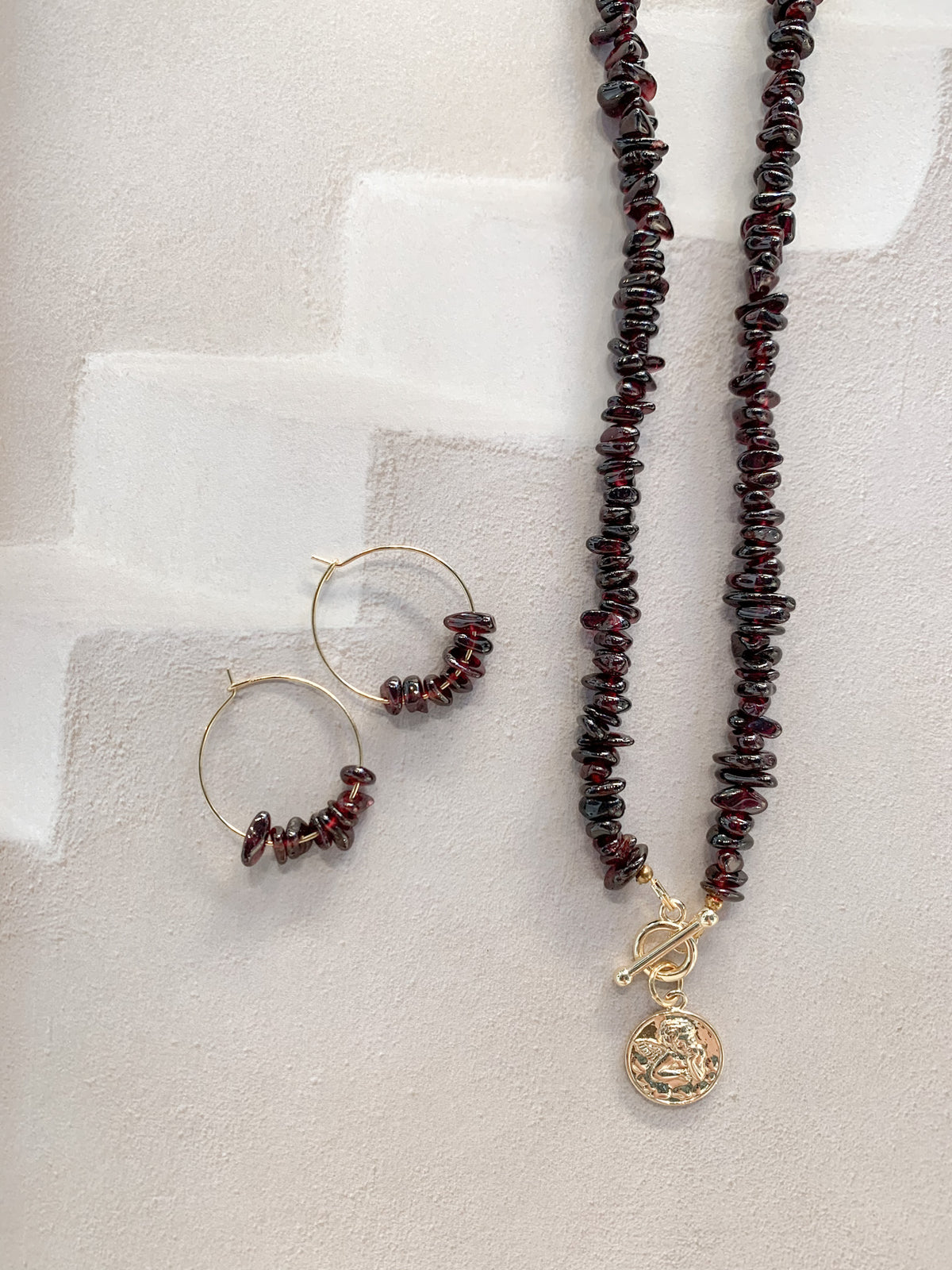 A jewelry set featuring a beaded necklace and matching hoop earrings. The necklace and hoops are made of dark reddish-brown garnet chip stones with gold-toned metal findings, including a toggle clasp and a round angel pendant, displayed on a textured light beige surface.