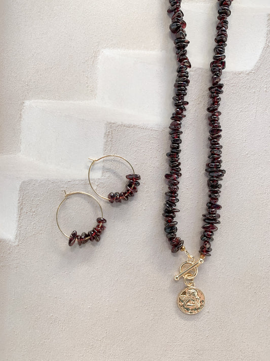 A jewelry set featuring a beaded necklace and matching hoop earrings. The necklace and hoops are made of dark reddish-brown garnet chip stones with gold-toned metal findings, including a toggle clasp and a round angel pendant, displayed on a textured light beige surface.