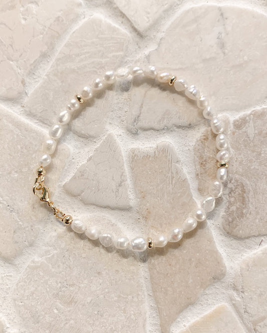 A delicate beaded bracelet with white freshwater pearls and small gold spacer beads, resting on a light beige stone tile background.