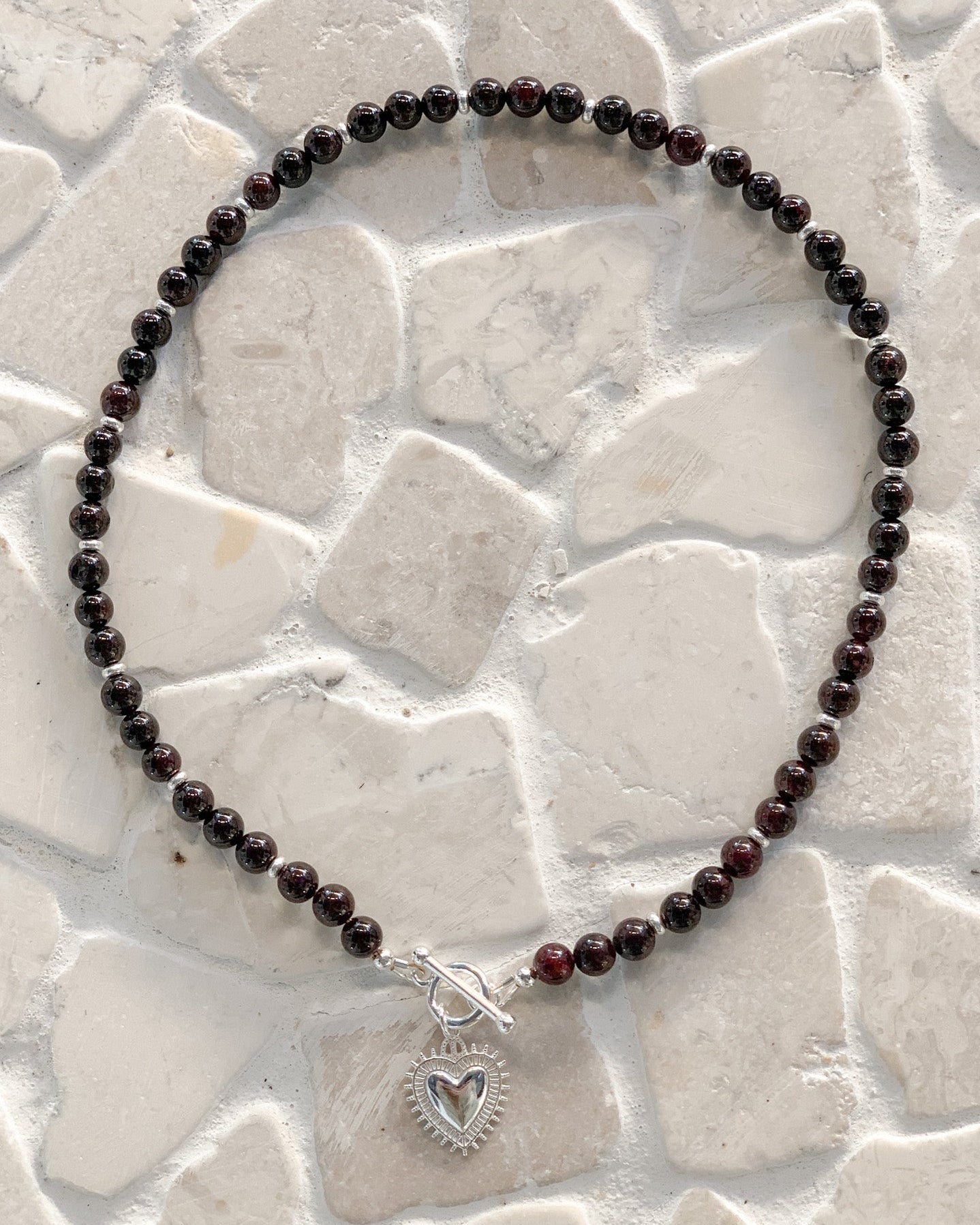 A beaded necklace made of round garnet beads, featuring a silver toggle clasp and a silver heart-shaped charm, displayed on a light beige stone tile background.