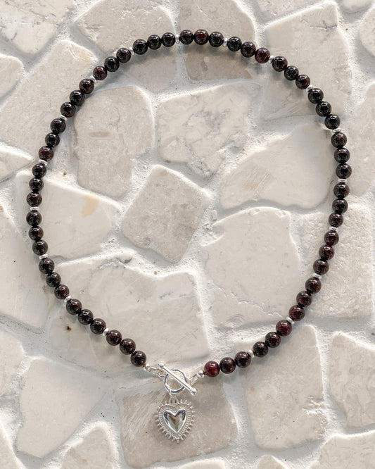 A beaded necklace made of round garnet beads, featuring a silver toggle clasp and a silver heart-shaped charm, displayed on a light beige stone tile background.