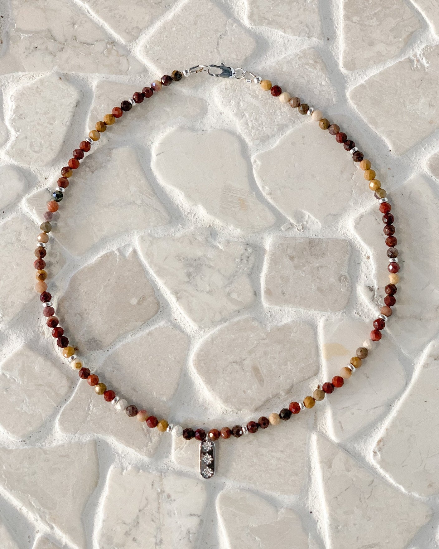 A beaded necklace with small Australian mookaite round beads in earthy red, brown, yellow, and white tones, featuring a silver-toned star pendant with cz stones, laid flat on a light stone-tile surface.
