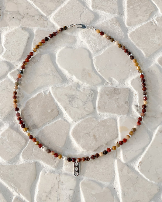 A beaded necklace with small Australian mookaite round beads in earthy red, brown, yellow, and white tones, featuring a silver-toned star pendant with cz stones, laid flat on a light stone-tile surface.