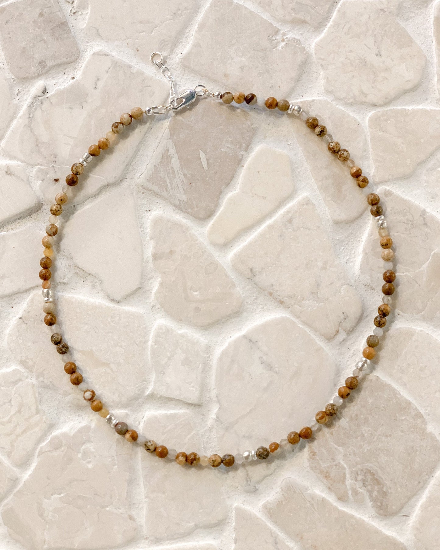 A beaded necklace made of small brown and tan picture jasper gemstone beads with a few clear faceted Botswana agate accents, laid in a circle on a light stone-tile surface.
