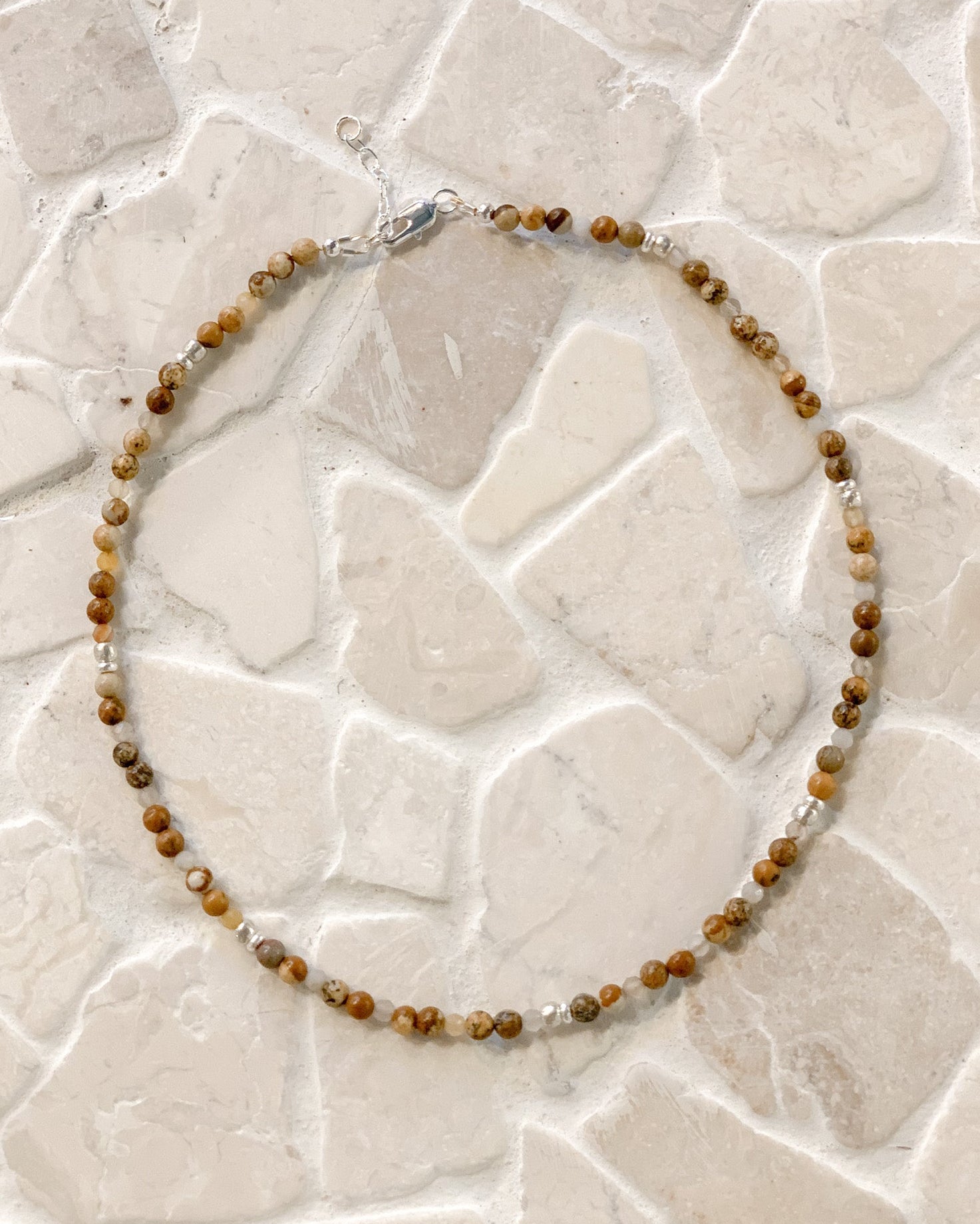 A beaded necklace made of small brown and tan picture jasper gemstone beads with a few clear faceted Botswana agate accents, laid in a circle on a light stone-tile surface.