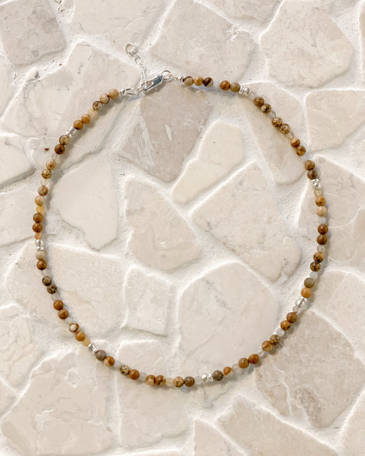 A beaded necklace made of small brown and tan picture jasper gemstone beads with a few clear faceted Botswana agate accents, laid in a circle on a light stone-tile surface.
