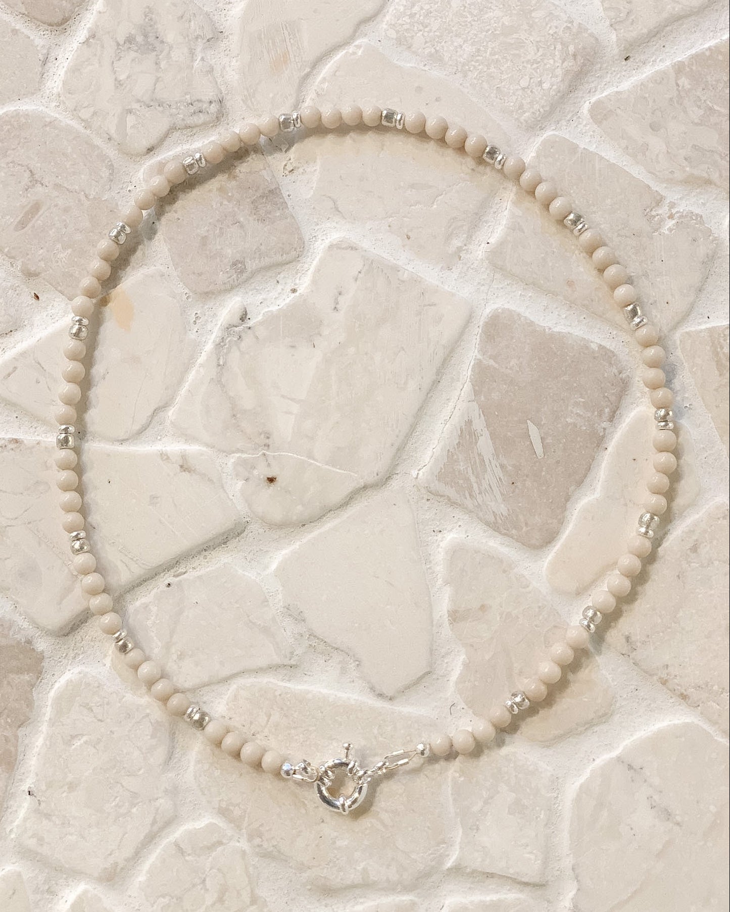 A beaded necklace made of small cream-colored Riverstone  beads with silver spacer accents and a silver clasp, laid flat on a light beige stone tile surface.