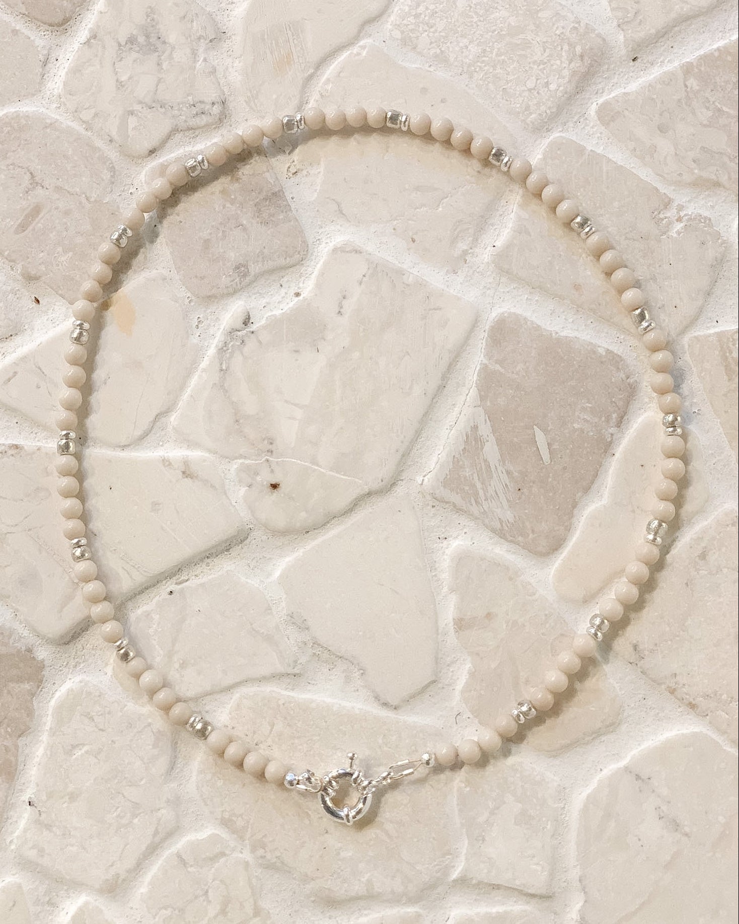 A beaded necklace made of small cream-colored Riverstone  beads with silver spacer accents and a silver clasp, laid flat on a light beige stone tile surface.