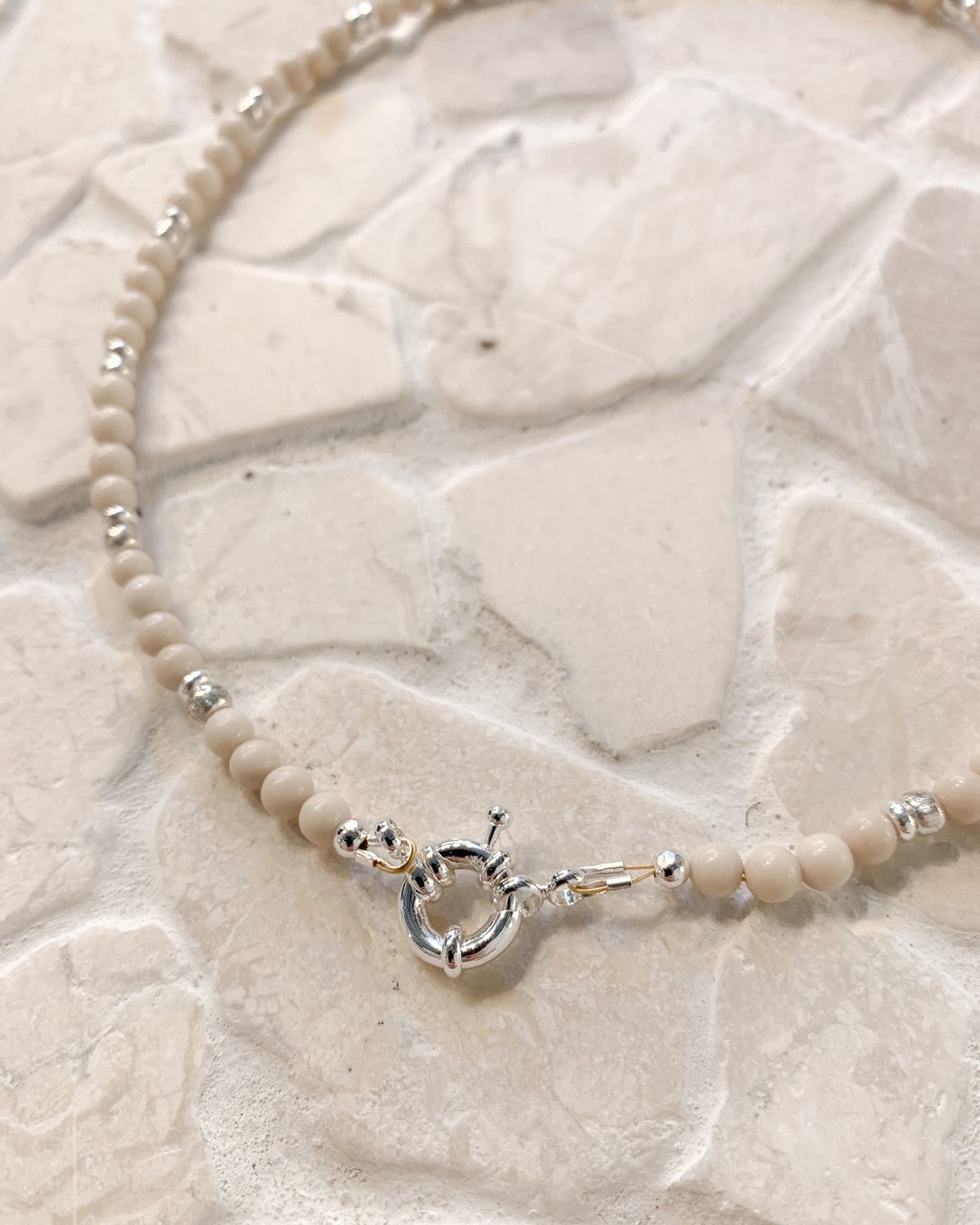 A delicate beaded necklace with small cream-colored Riverstone beads and silver spacer accents, featuring a silver toggle clasp, displayed on a textured light beige stone surface.