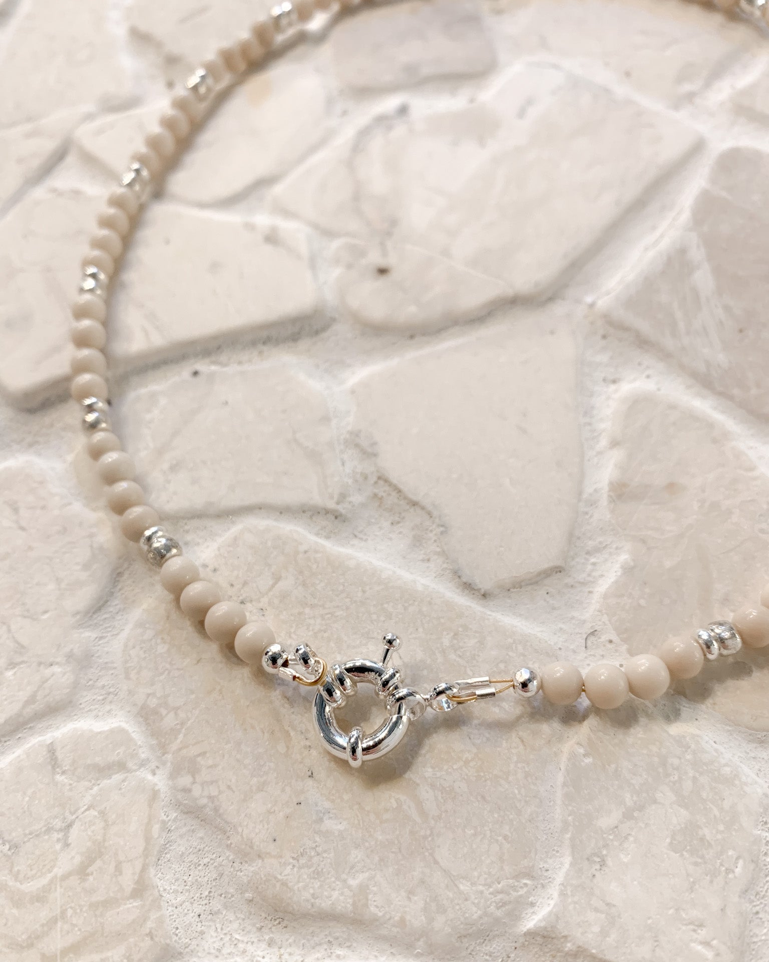 A delicate beaded necklace with small cream-colored Riverstone beads and silver spacer accents, featuring a silver toggle clasp, displayed on a textured light beige stone surface.