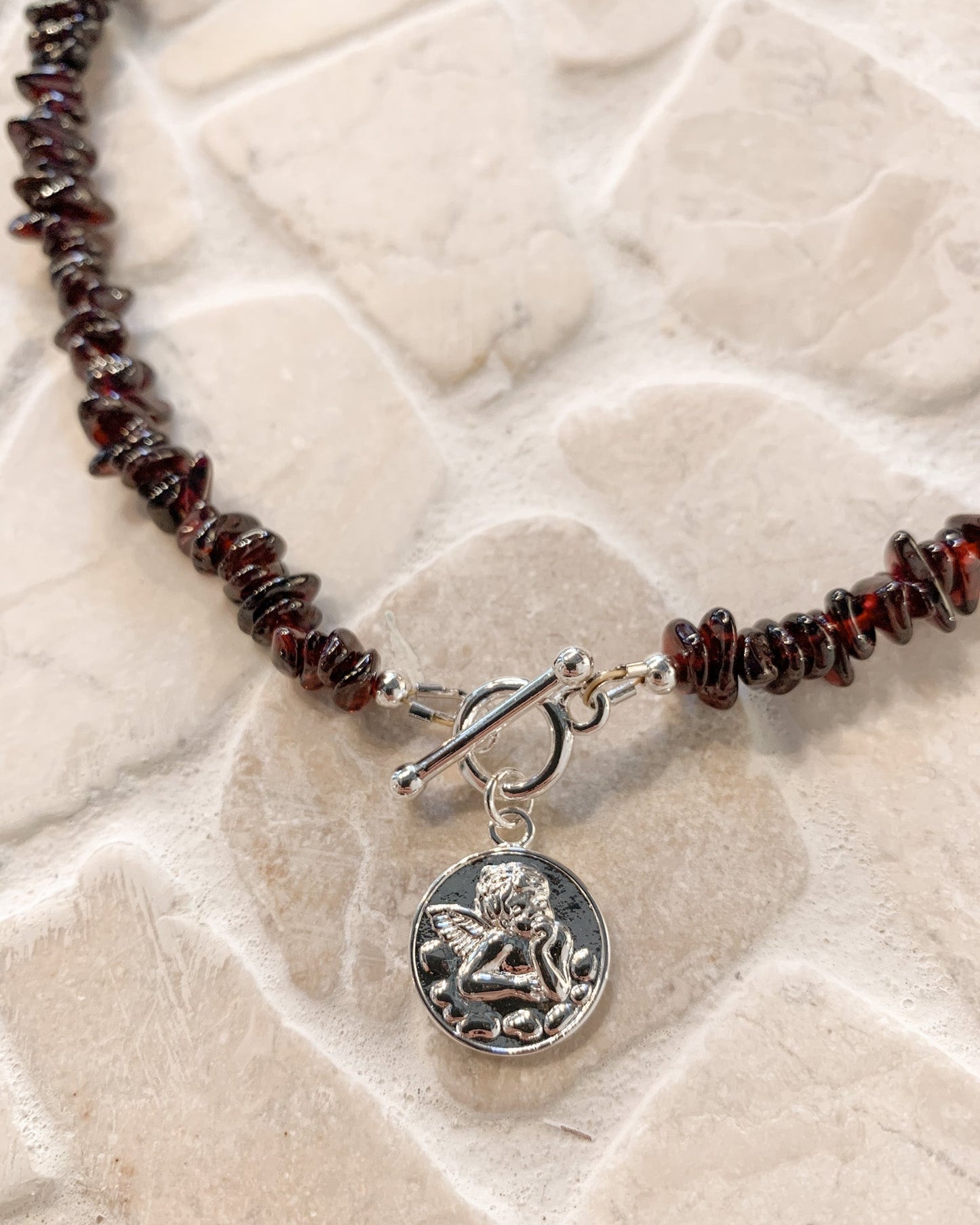 A close-up of a beaded necklace with dark reddish-brown polished garnet gemstone beads, a silver toggle clasp, and a round silver charm featuring an embossed cherub angel, displayed on a light beige stone surface.