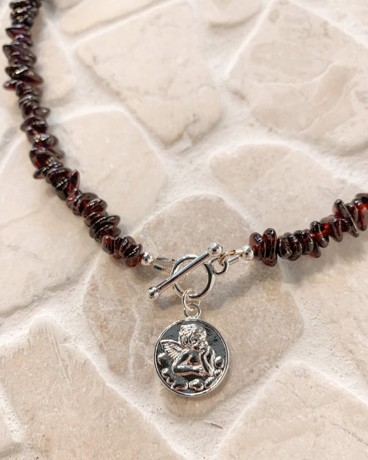 A close-up of a beaded necklace with dark reddish-brown polished garnet gemstone beads, a silver toggle clasp, and a round silver charm featuring an embossed cherub angel, displayed on a light beige stone surface.