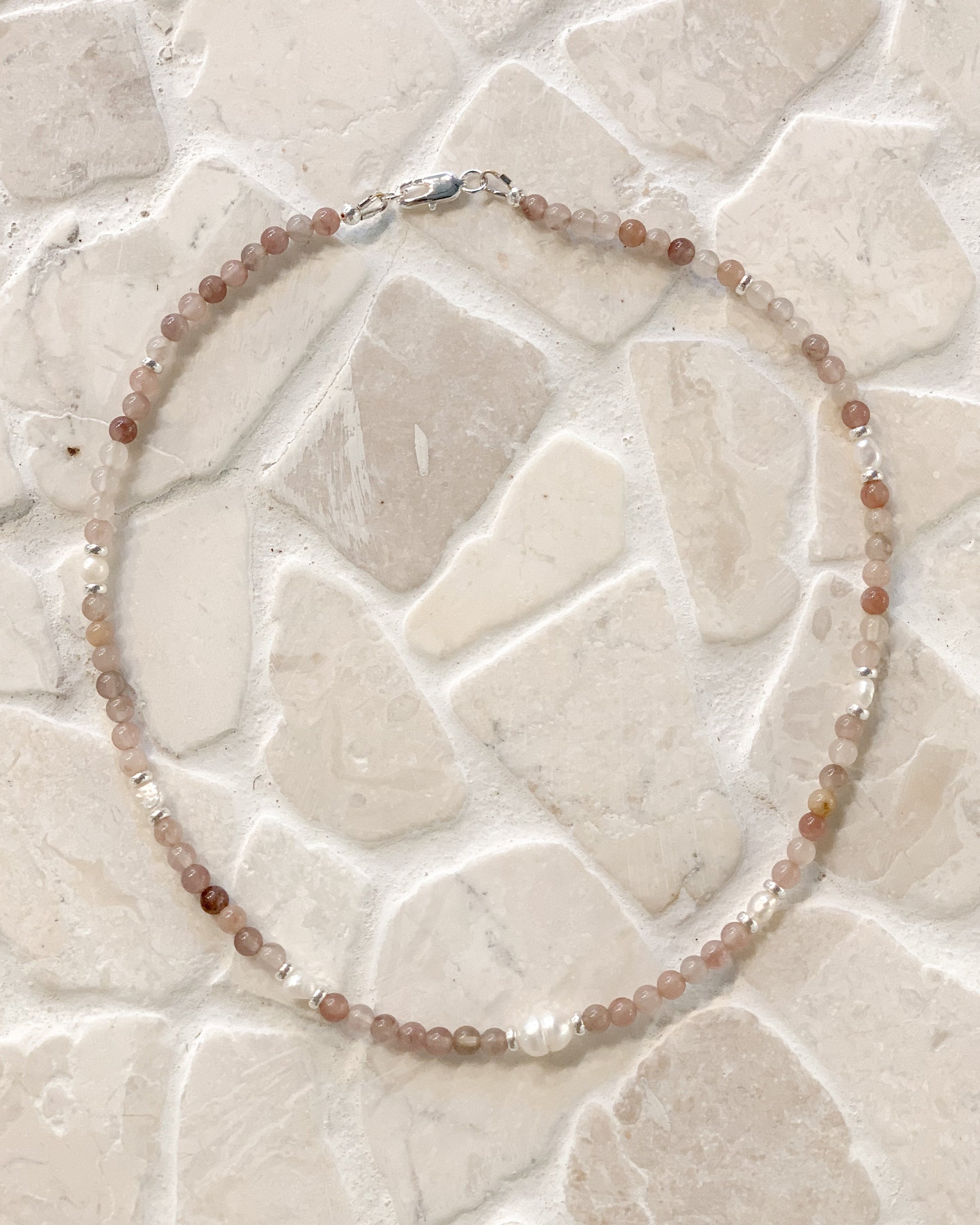 A beaded necklace made of small translucent peach, pink, and brown beads with a few clear faceted beads and a silver clasp, laid flat on a light beige stone tile background.