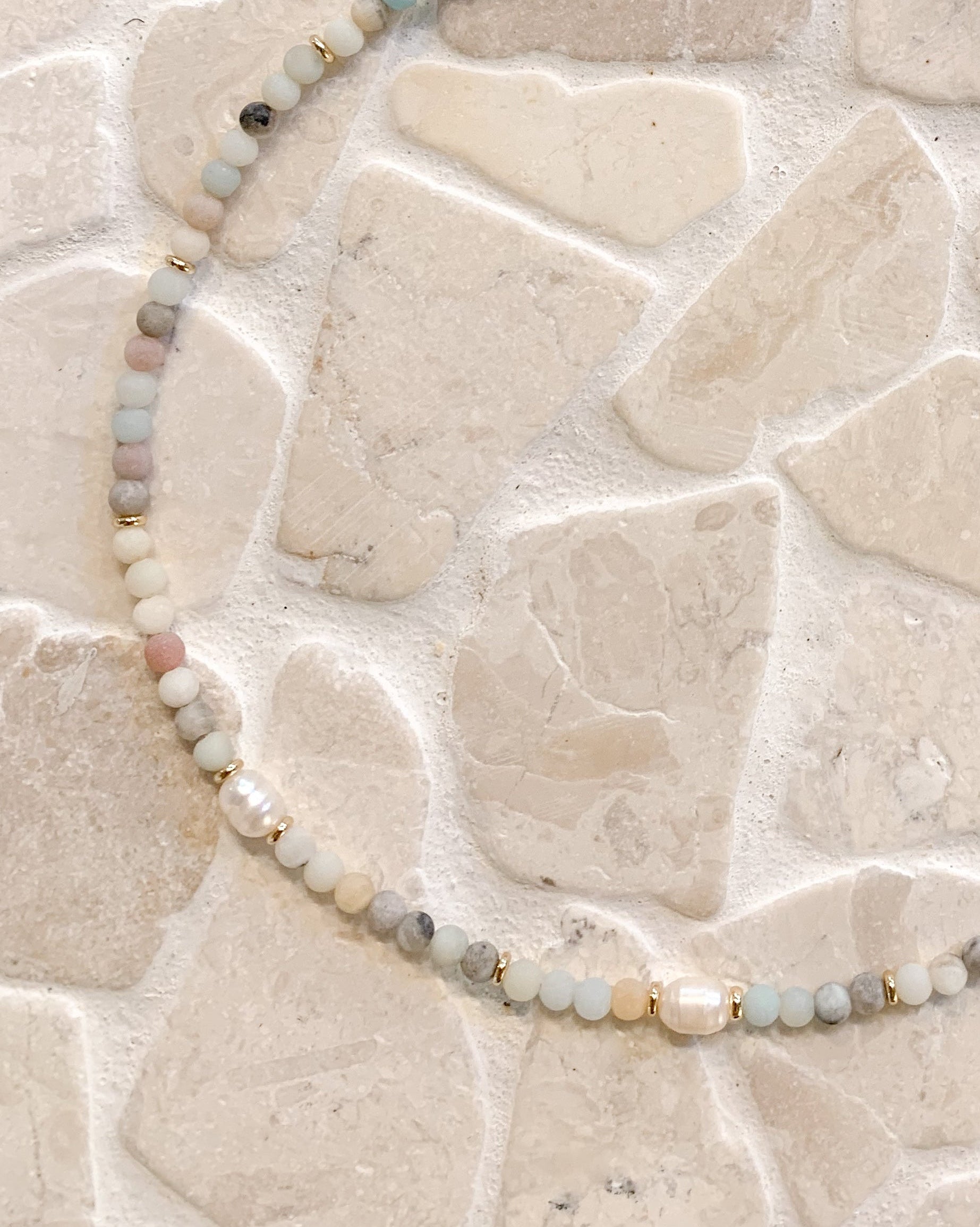 A beaded necklace made of small ammonite beads and pearls, accented by tiny gold spacers and a gold clasp, displayed on a light beige stone tile background.