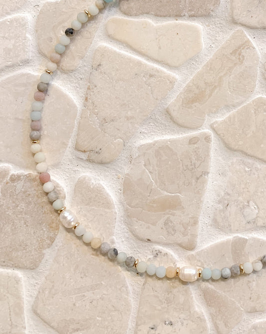 A beaded necklace made of small ammonite beads and pearls, accented by tiny gold spacers and a gold clasp, displayed on a light beige stone tile background.