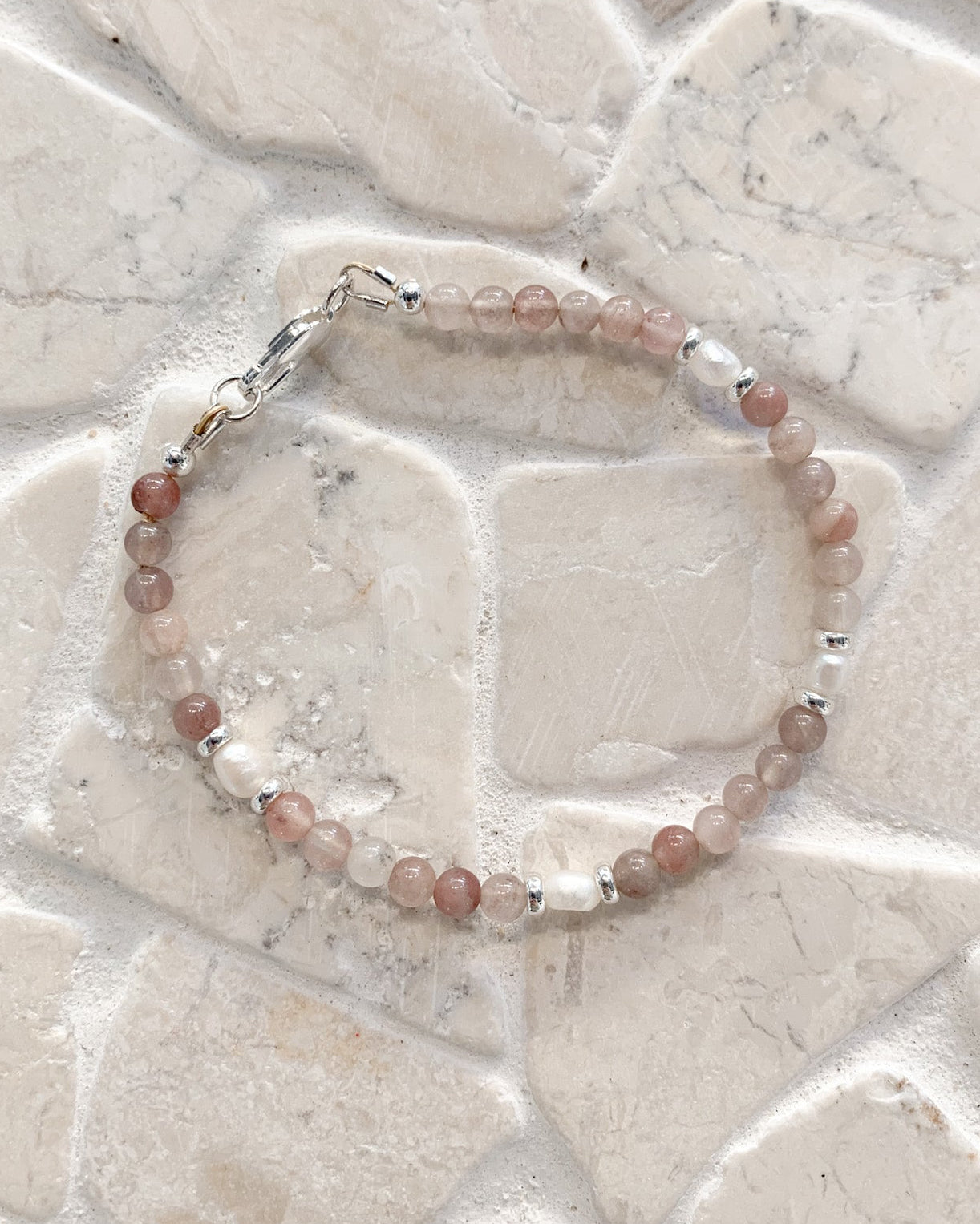A gemstone and pearl beaded bracelet made of dusty pink and light brown translucent beads with small silver spacer beads and a silver clasp, displayed on a light beige stone tile background.