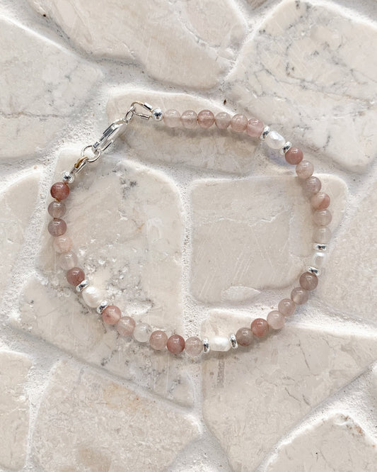A gemstone and pearl beaded bracelet made of dusty pink and light brown translucent beads with small silver spacer beads and a silver clasp, displayed on a light beige stone tile background.