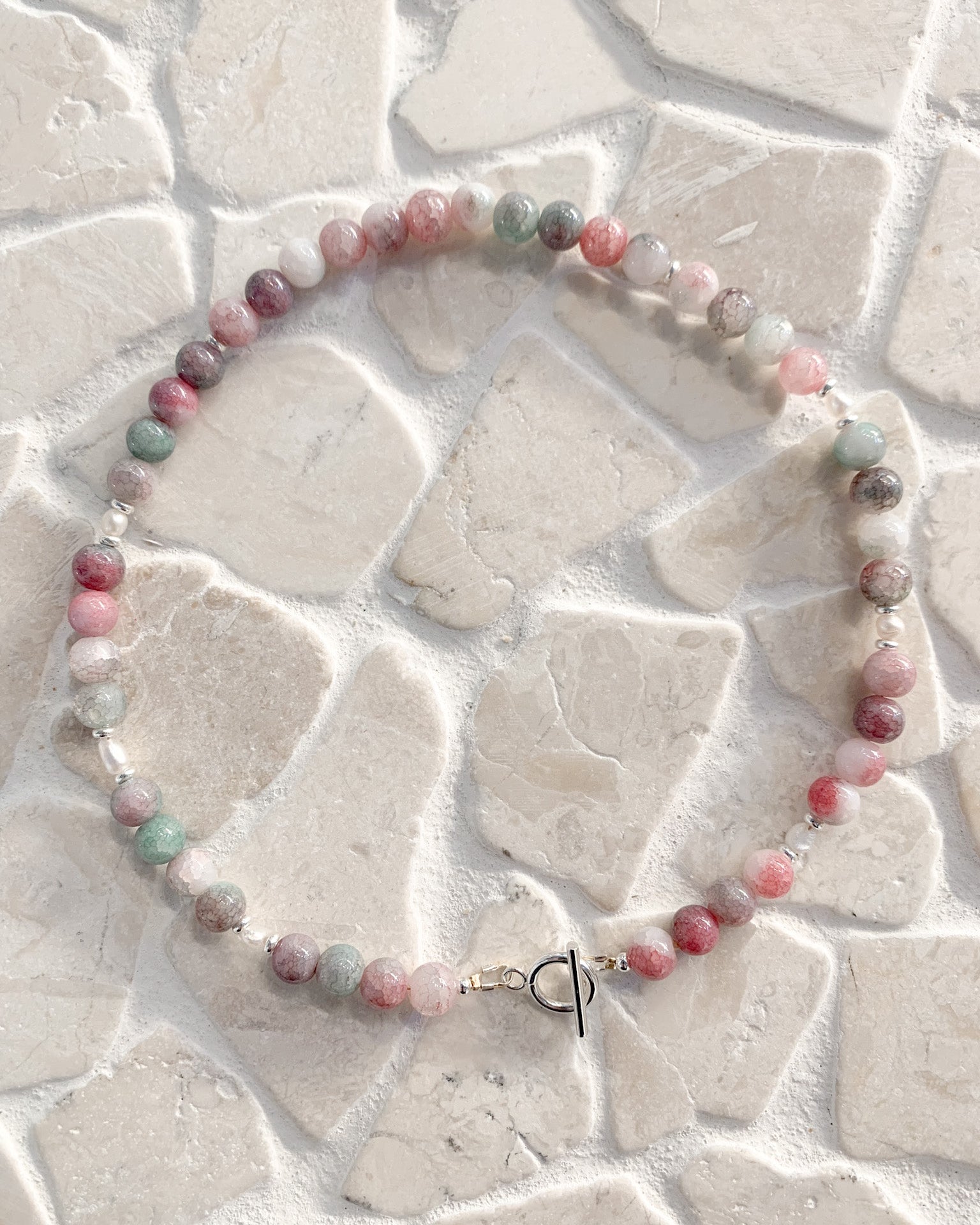 A beaded necklace made of round, polished gemstone beads in pink, green, gray, and white, arranged in a circle on a light beige stone tile background. The necklace features a small silver toggle clasp.