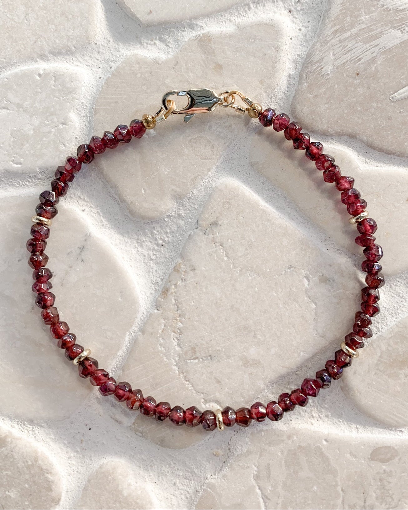 A beaded bracelet made of faceted dark red garnet beads with small gold spacer beads and a gold clasp, photographed on a light stone surface.