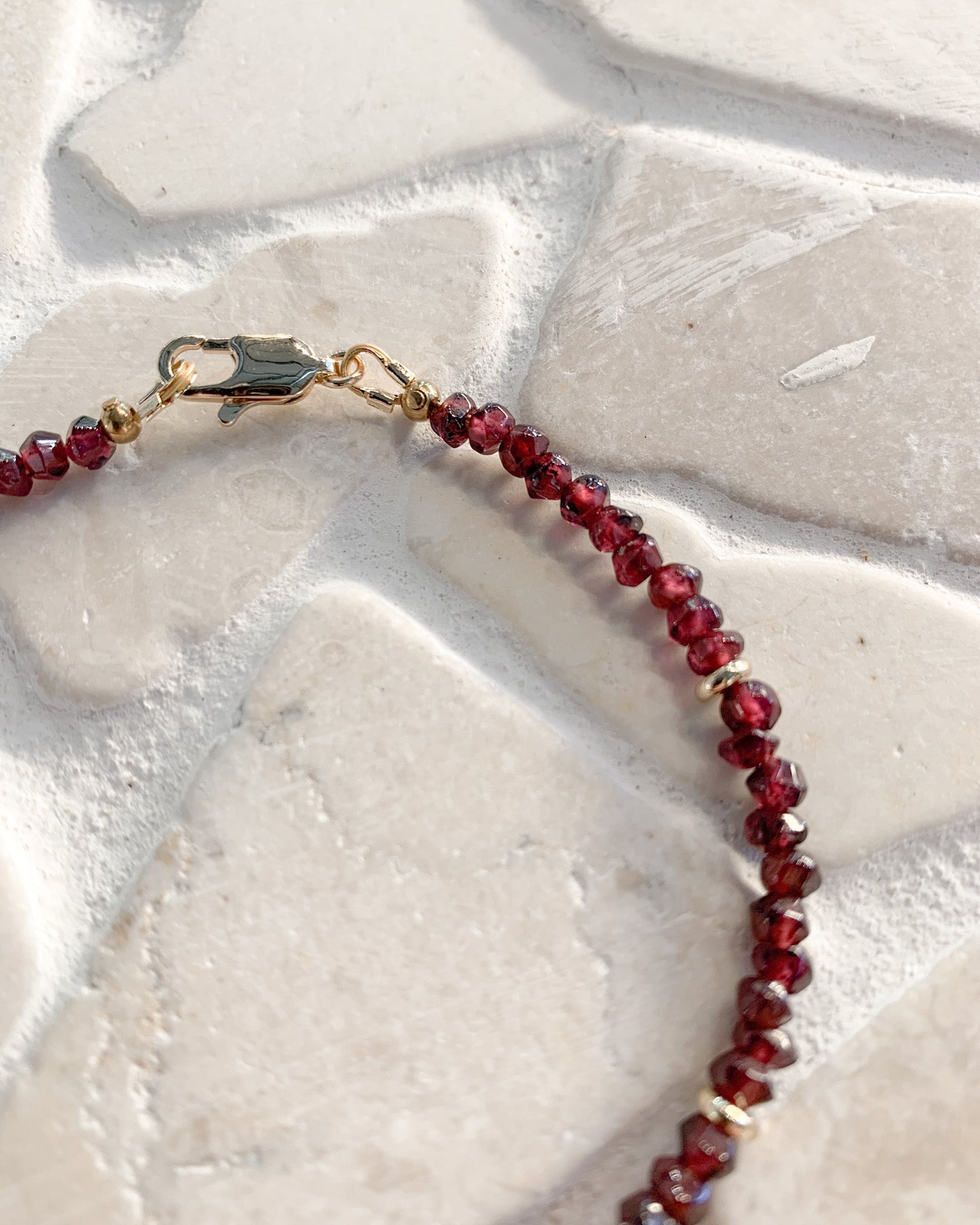 A close-up of a beaded necklace with faceted dark red garnet gemstone beads and small gold spacer rings, attached to a gold lobster clasp, laid on a light beige stone surface.