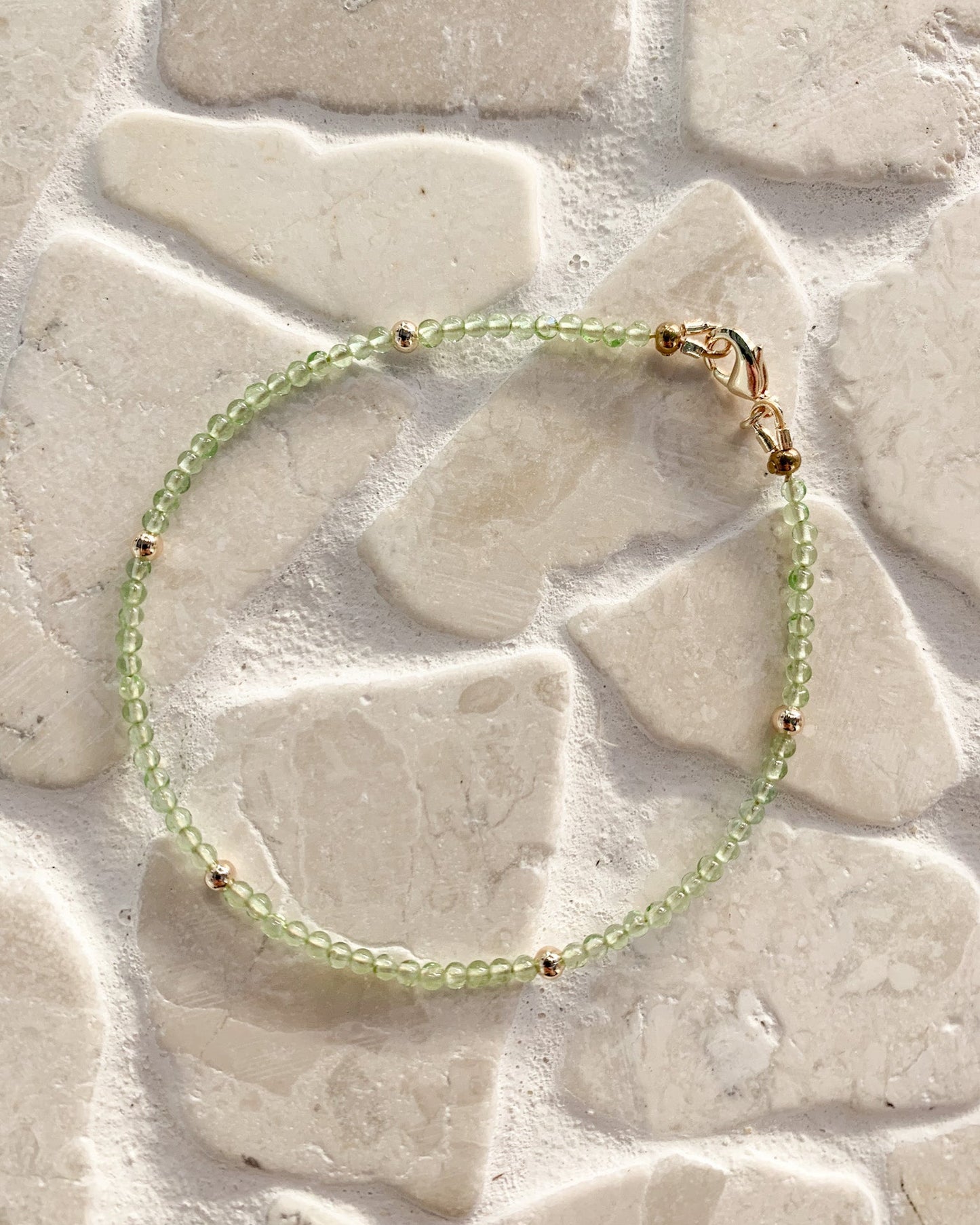 A delicate beaded bracelet with light green translucent peridot beads and small gold spacer beads, resting on a light beige stone tile surface.
