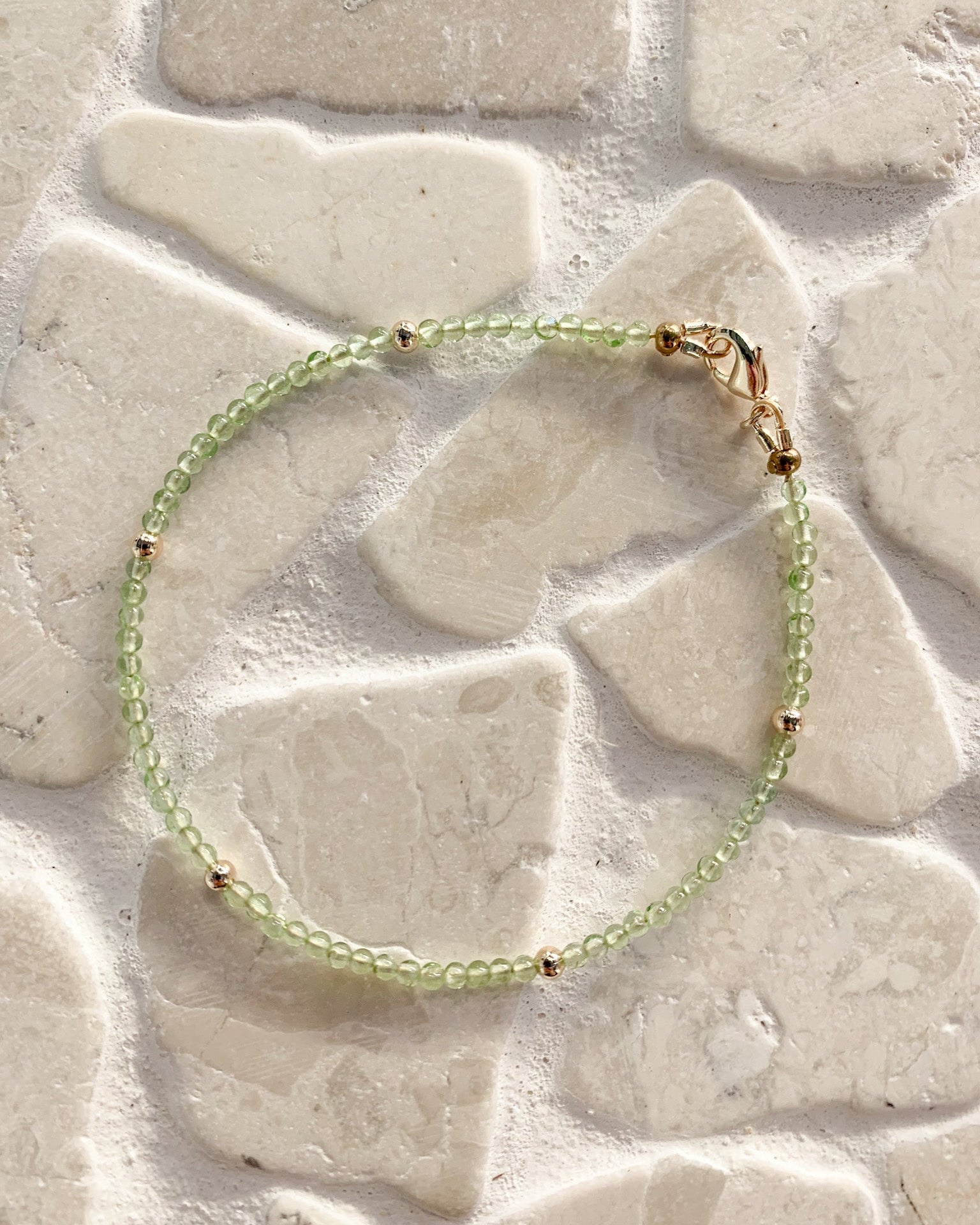 A delicate beaded bracelet with light green translucent peridot beads and small gold spacer beads, resting on a light beige stone tile surface.
