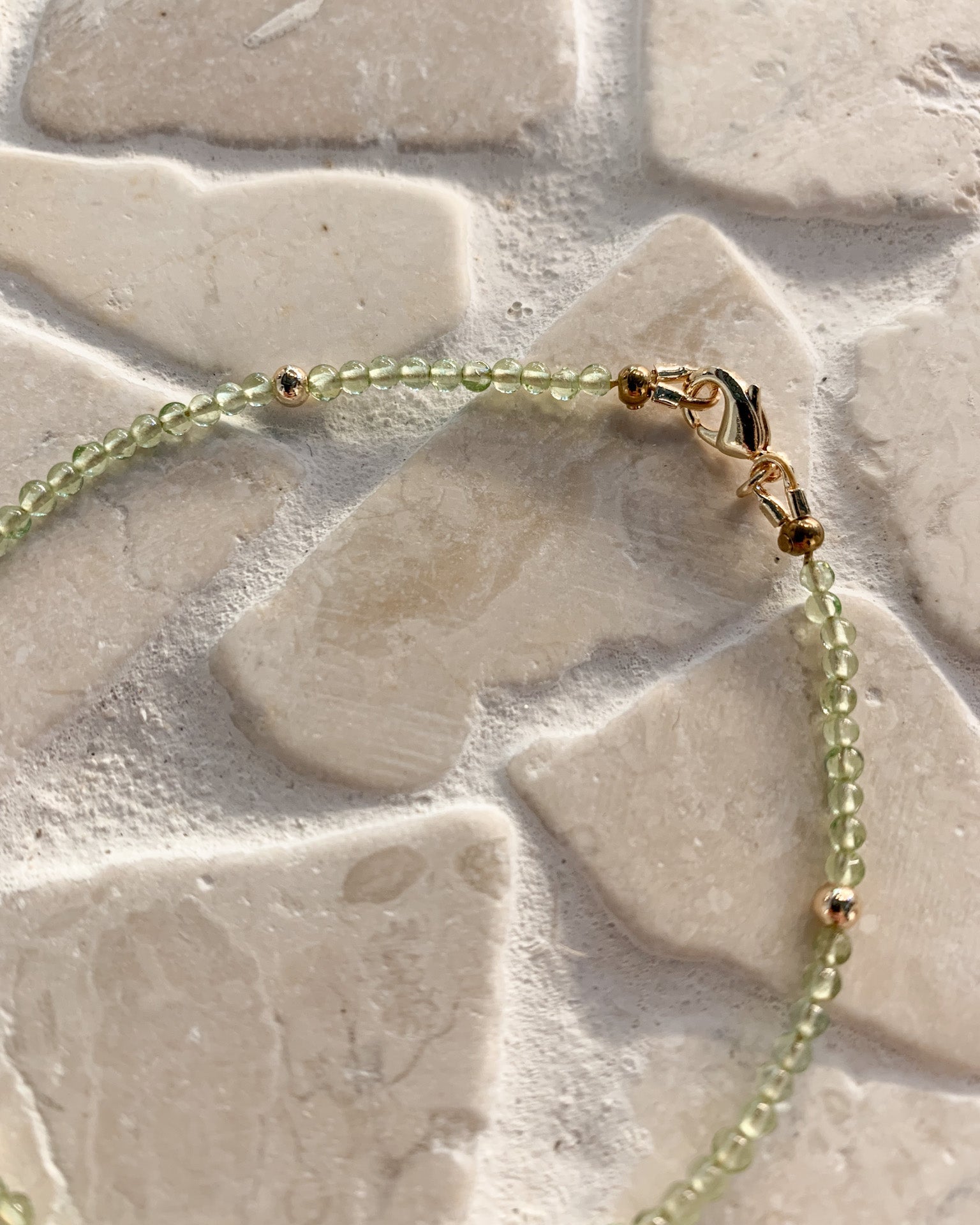 A delicate beaded bracelet with light green peridot beads and small gold spacer beads, featuring a gold lobster clasp, laid out on a light beige stone tile surface.