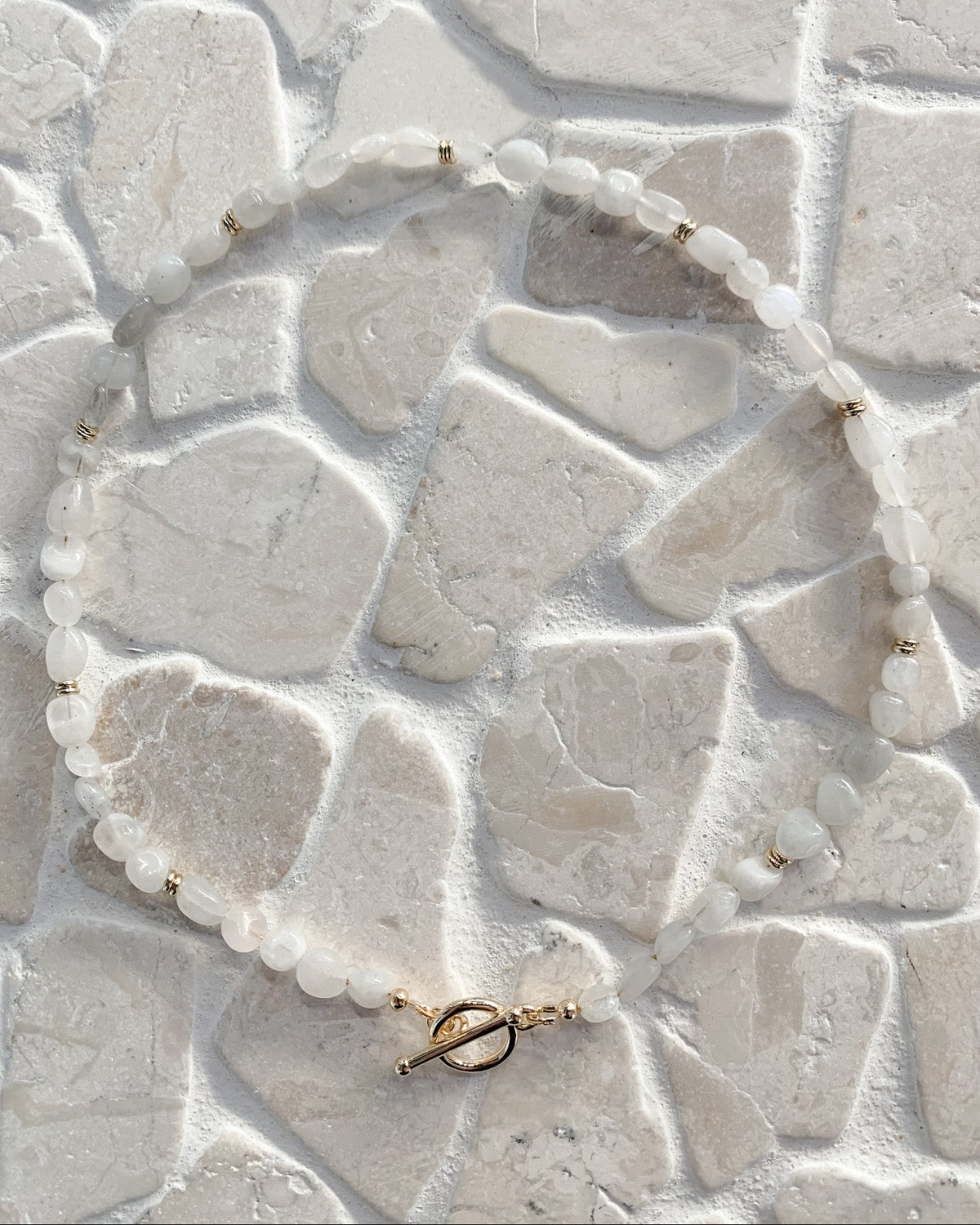 A beaded necklace made of round white moonstone gemstone beads separated by small gold spacers, with a gold toggle clasp, laid out on a light stone tile surface.