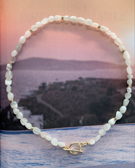 A beaded necklace made of irregular white moonstone beads with small gold spacer accents and a gold toggle clasp, displayed against a scenic background featuring a pink sky and blue water.